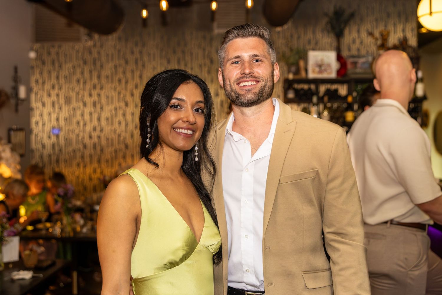 A smiling couple poses together at an indoor event, woman in green dress, man in tan blazer.