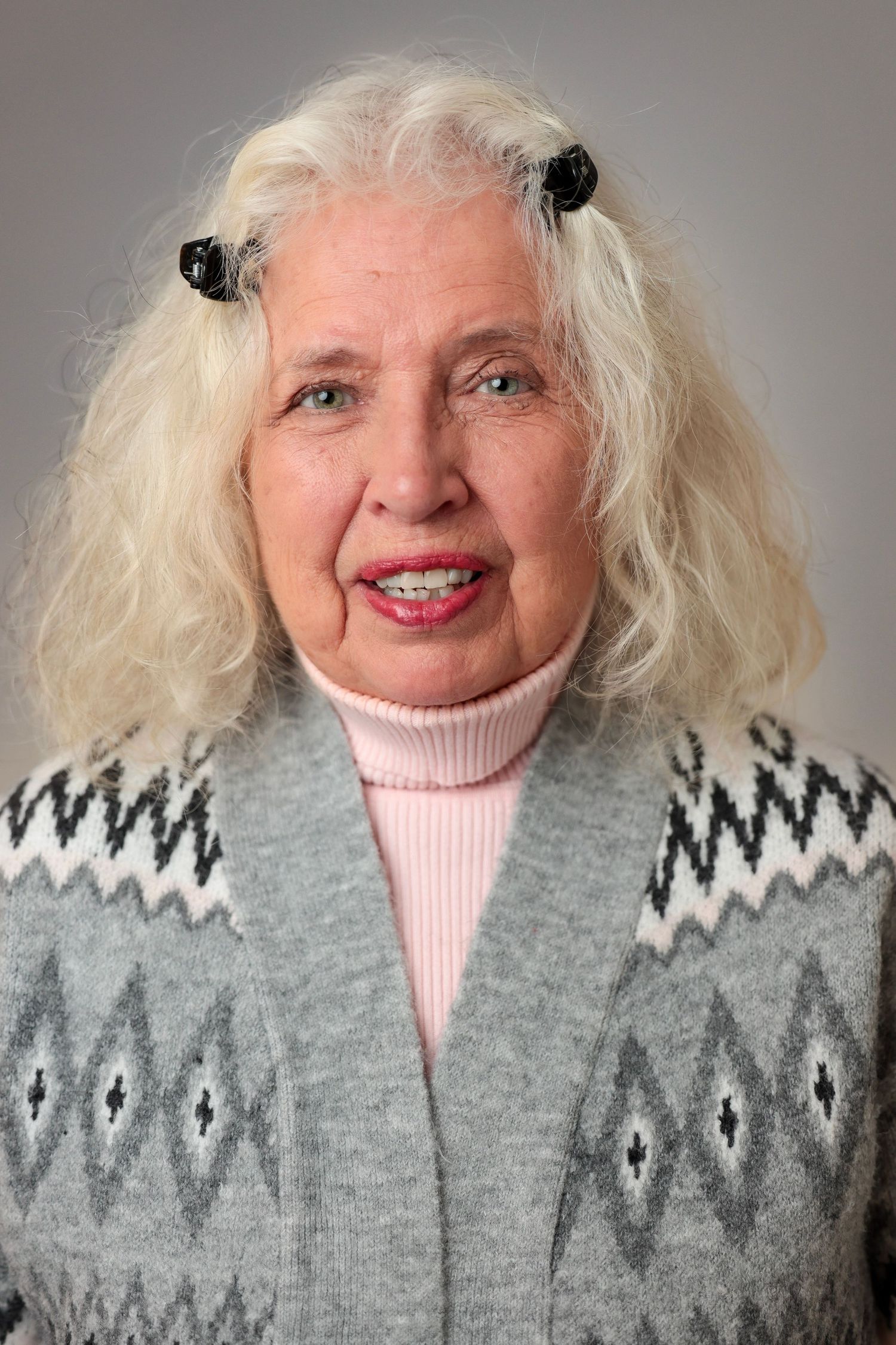 Senior woman in gray patterned sweater and pink turtleneck smiling warmly at camera.