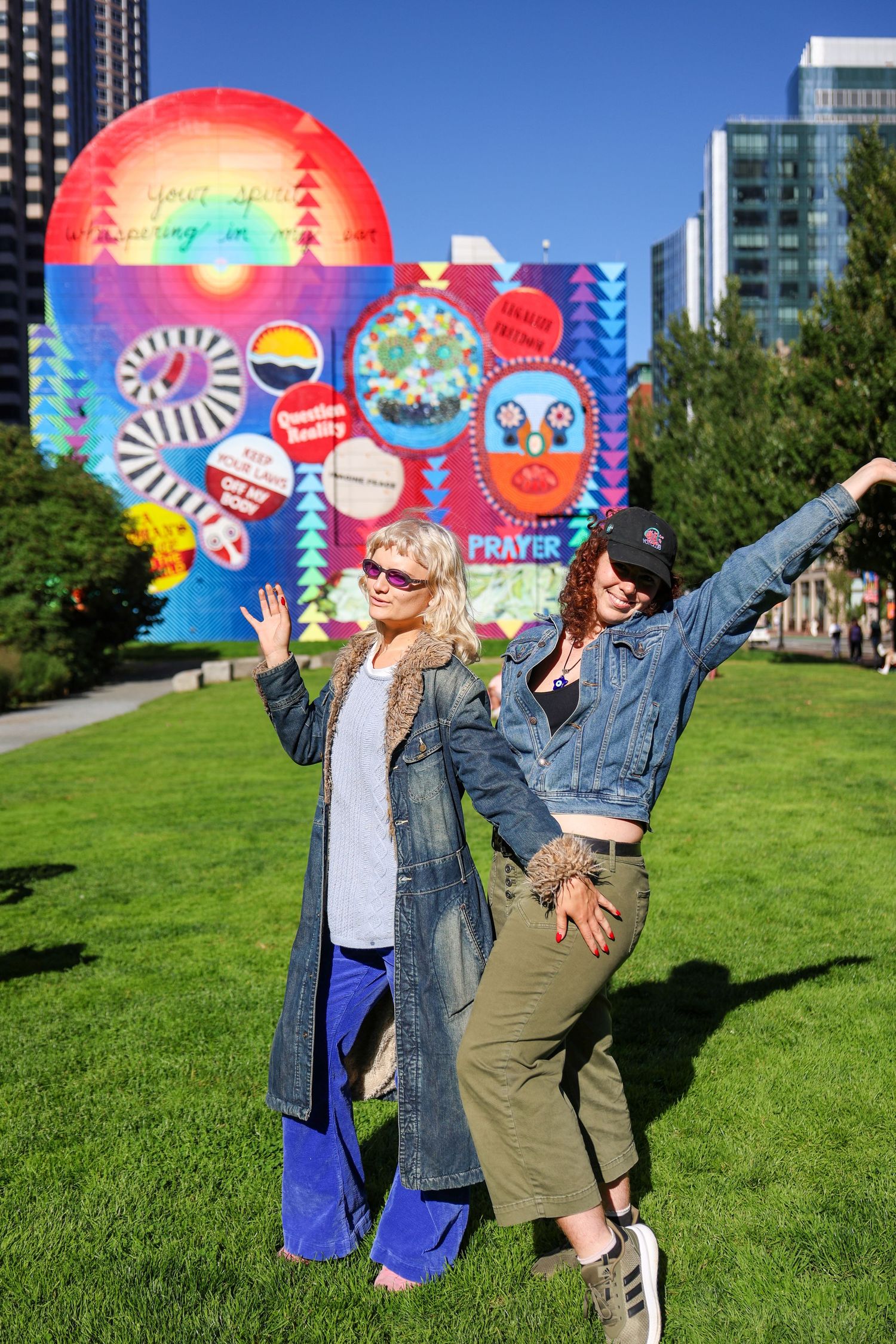 Playful poses in front of bright street art wall on grassy lawn with city buildings in background.
