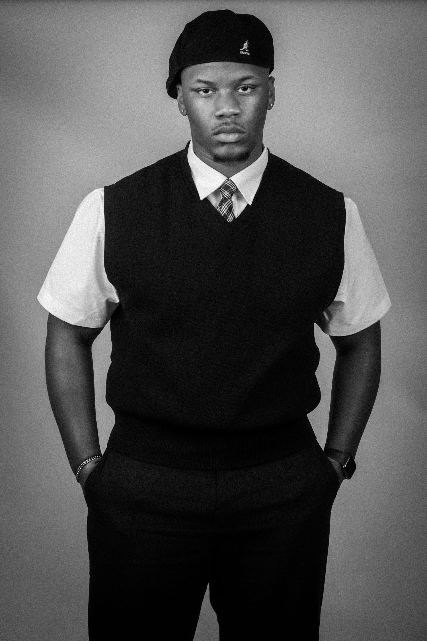 Black and white portrait of a young man in a vest, dress shirt, tie, and cap, posing confidently.