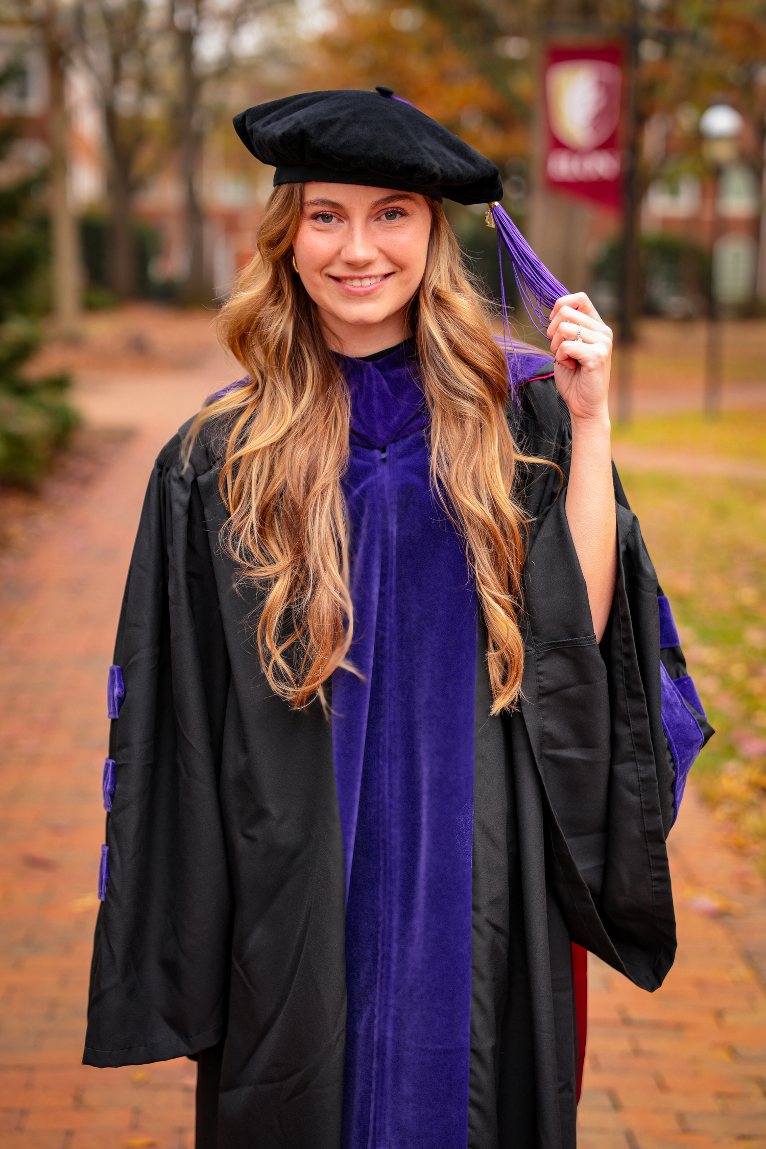 Graduate wearing black cap and purple academic regalia poses on brick pathway during autumn graduation ceremony.