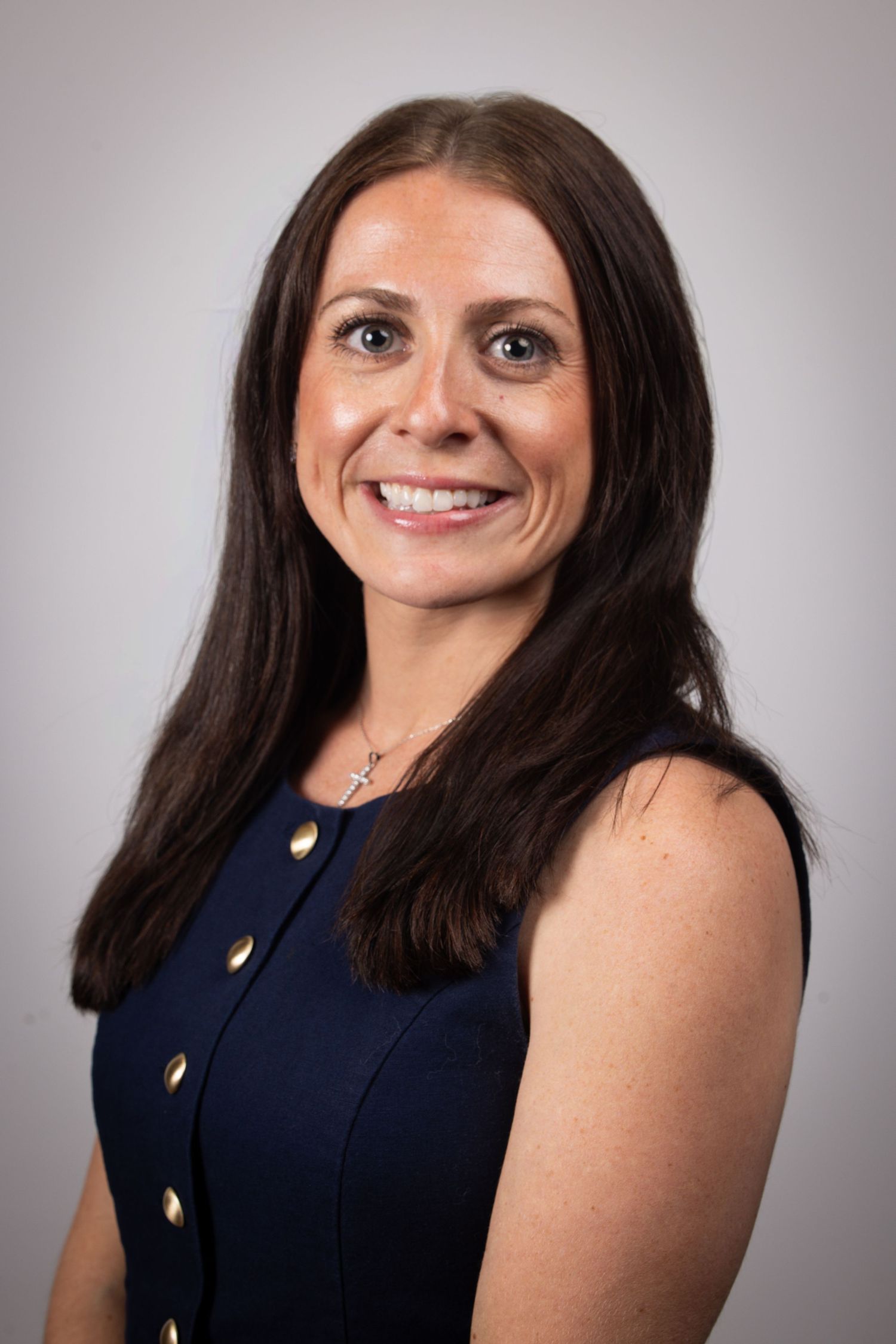Professional headshot of a smiling woman with long dark hair wearing a navy sleeveless dress with gold buttons and cross necklace.