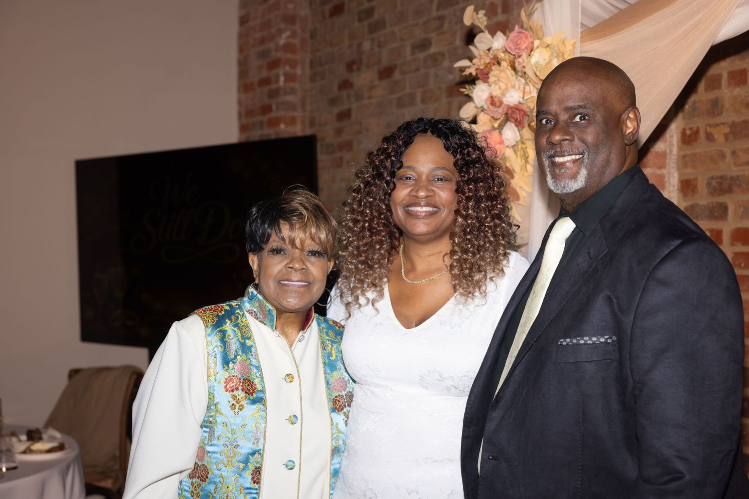 Three people smiling and posing together at a formal event in an elegant venue with brick walls.