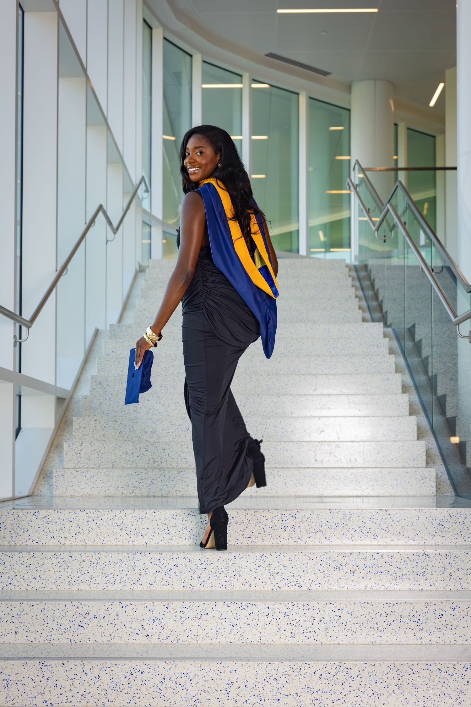A smiling Black woman in a black dress walks up stairs wearing blue and gold graduation regalia, holding a cap.
