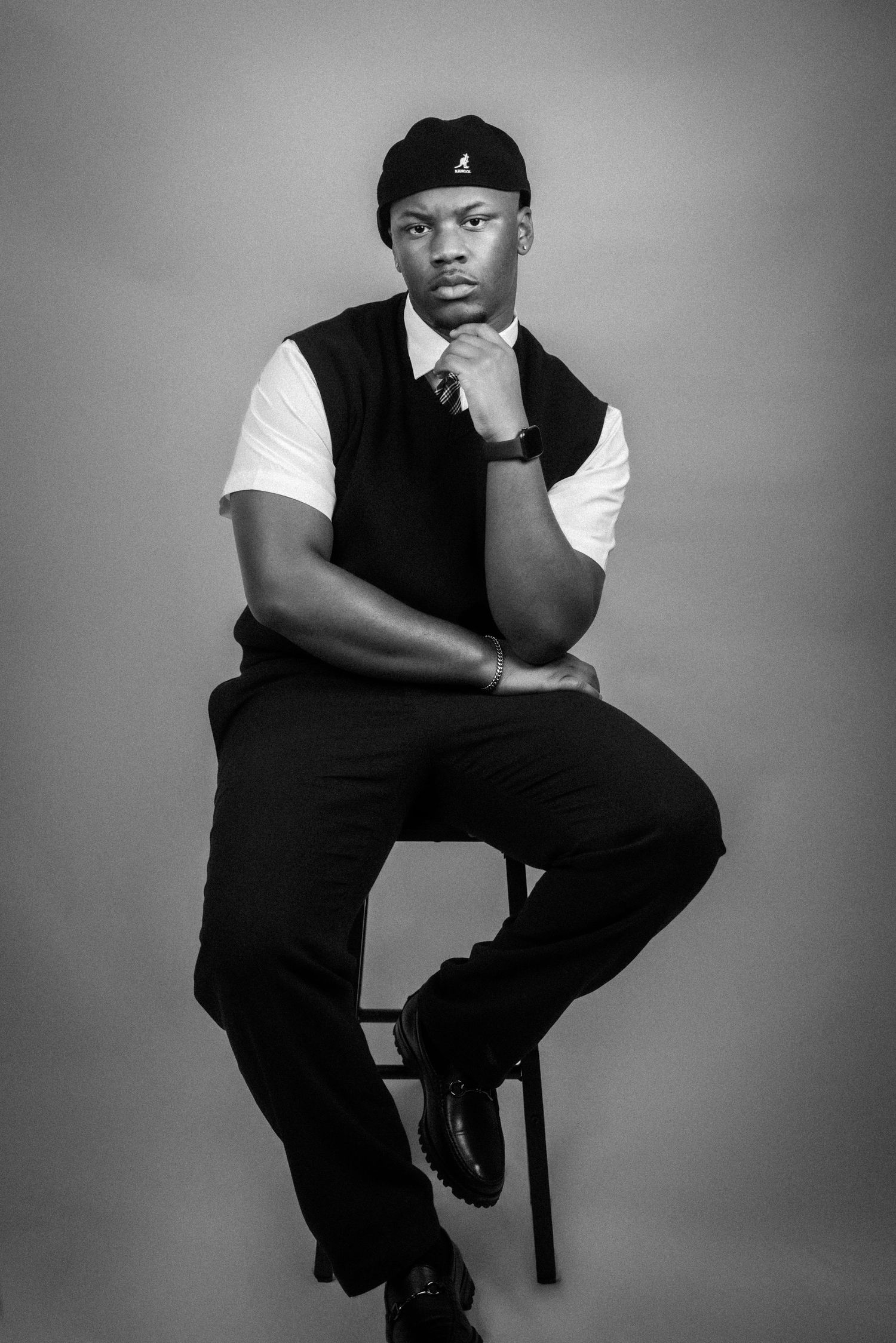 Black and white portrait of a young man in vest and cap seated on stool with hand on chin.
