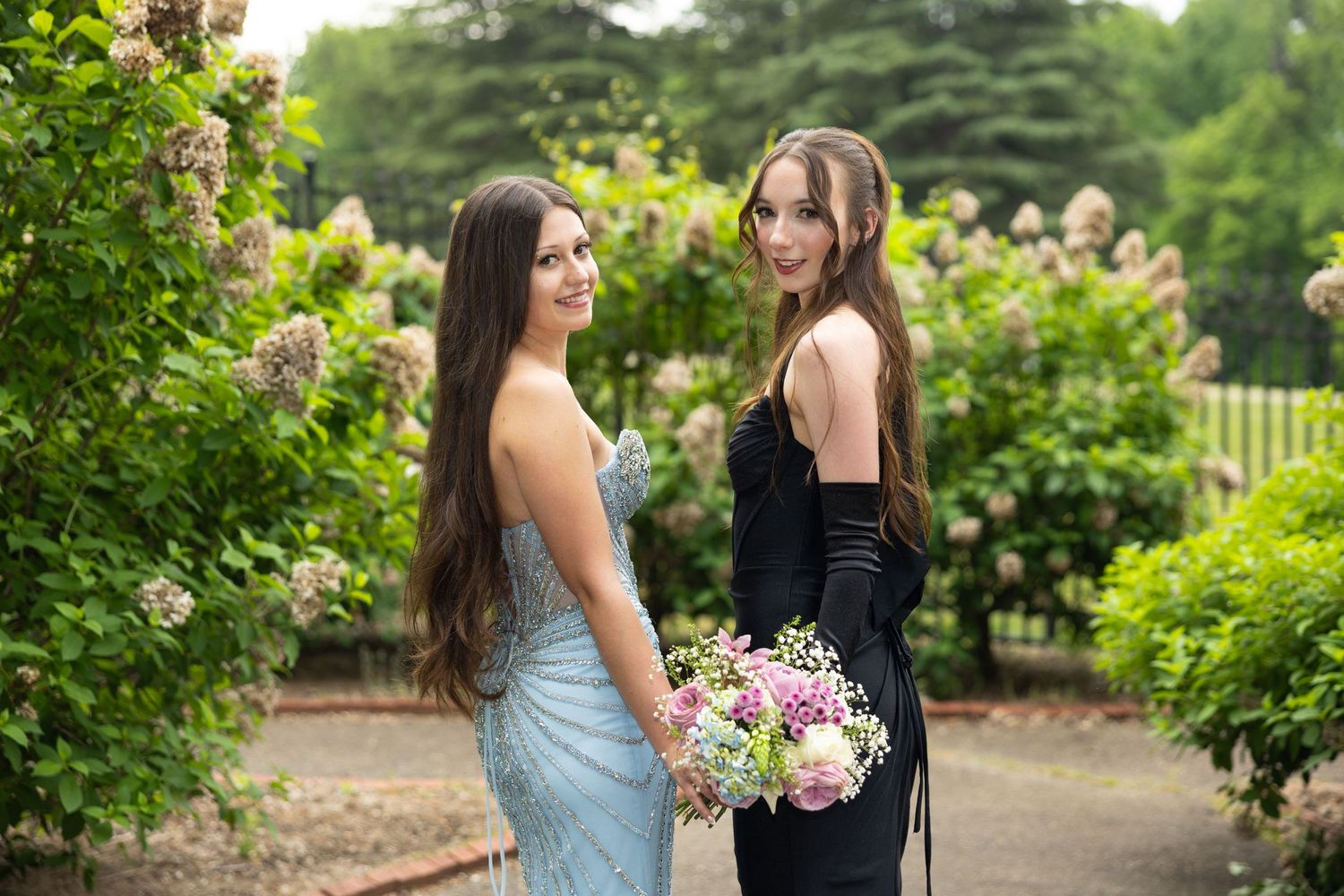 Two elegant young women in formal gowns pose in a lush garden, one holding a bouquet of pink flowers.
