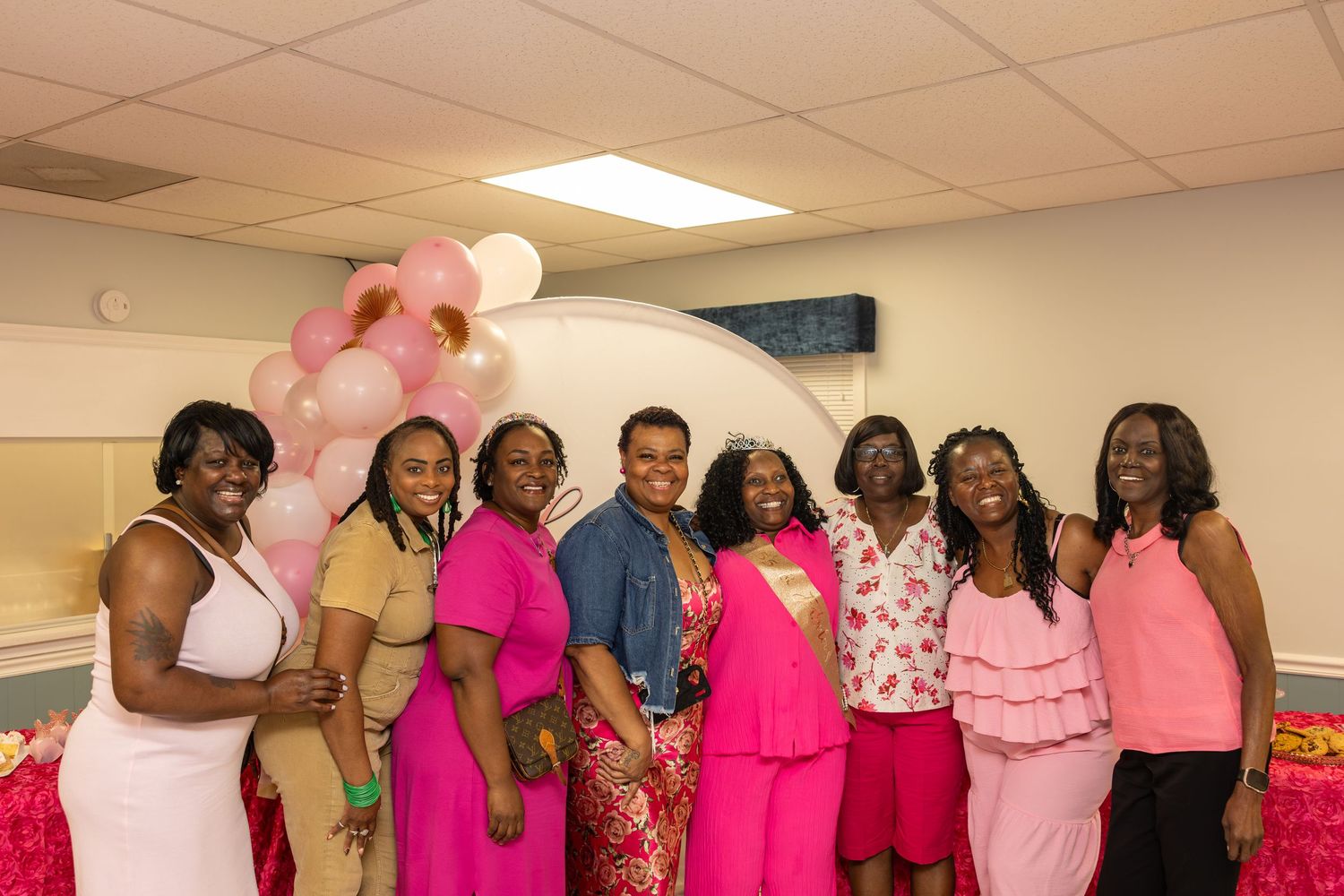 Group of nine women in pink outfits smiling together at a pink-themed celebration event with balloon decorations.