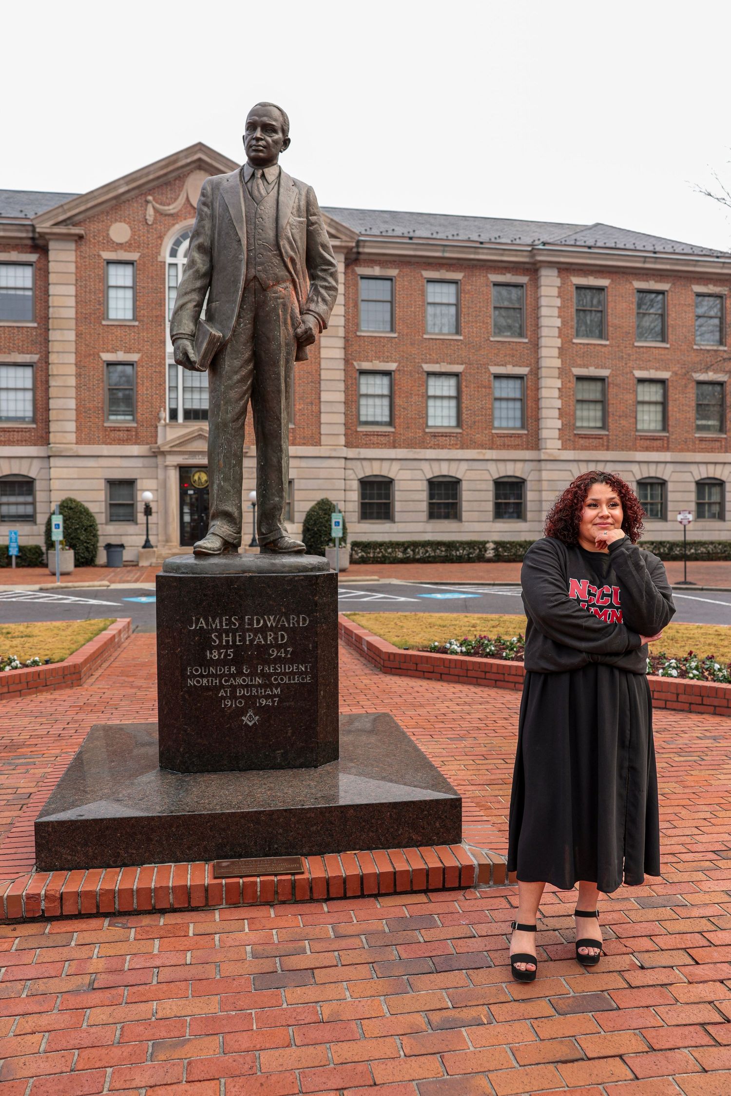 Bronze statue memorial on a brick plaza in front of a historic brick building with decorative landscaping.