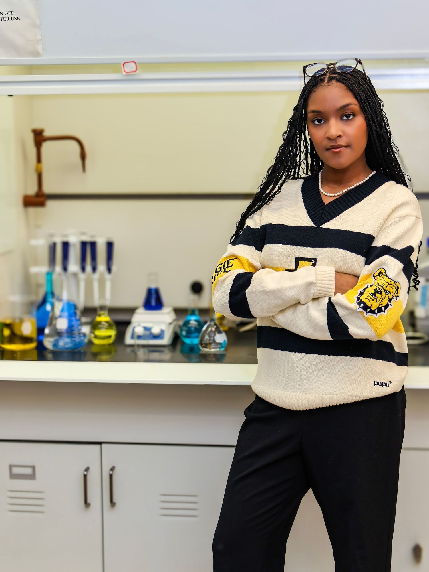 Student wearing black and cream university sweatshirt poses in science laboratory with equipment.