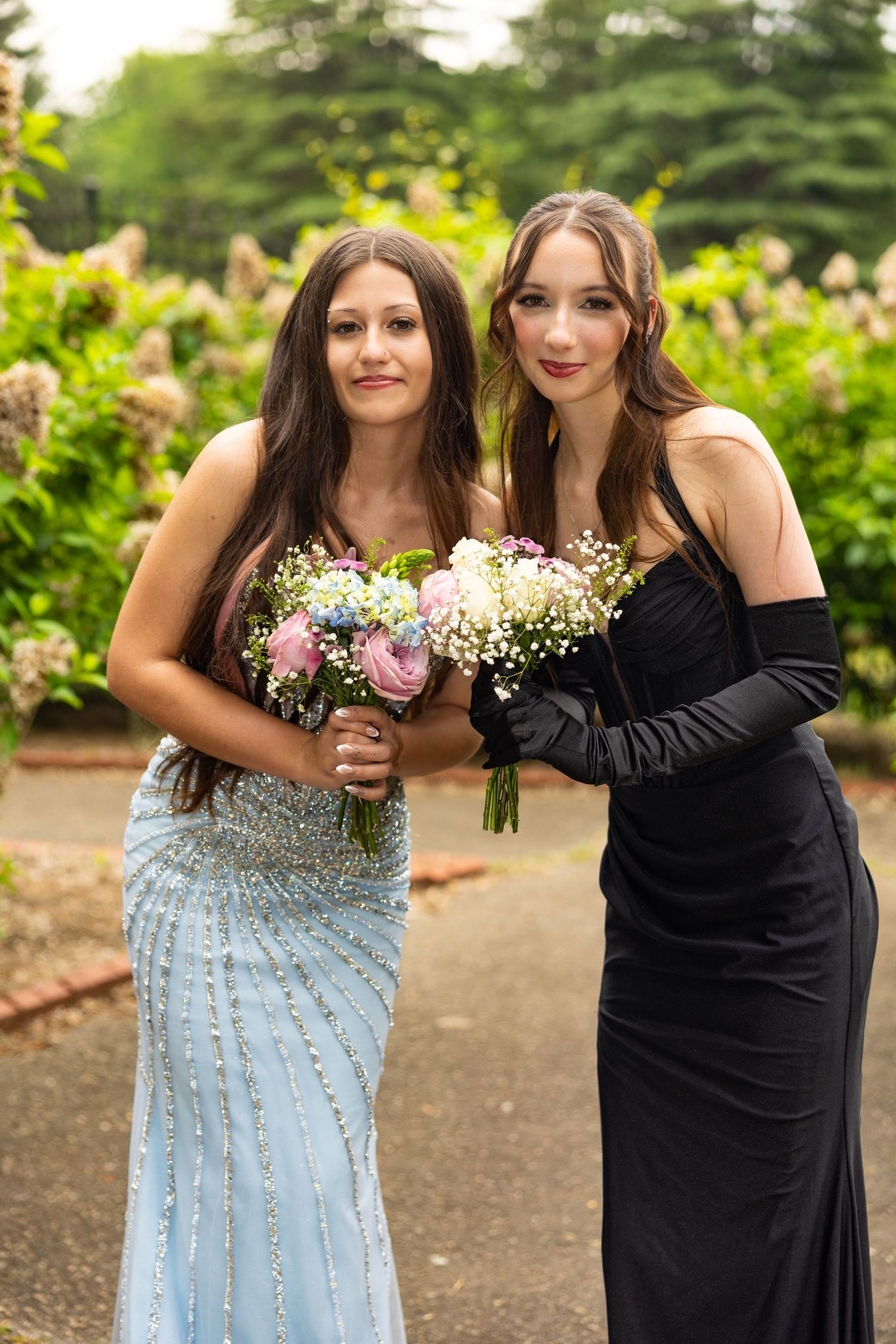 Two young women in elegant prom gowns holding floral bouquets, one in blue sequined dress, one in black gown with gloves.