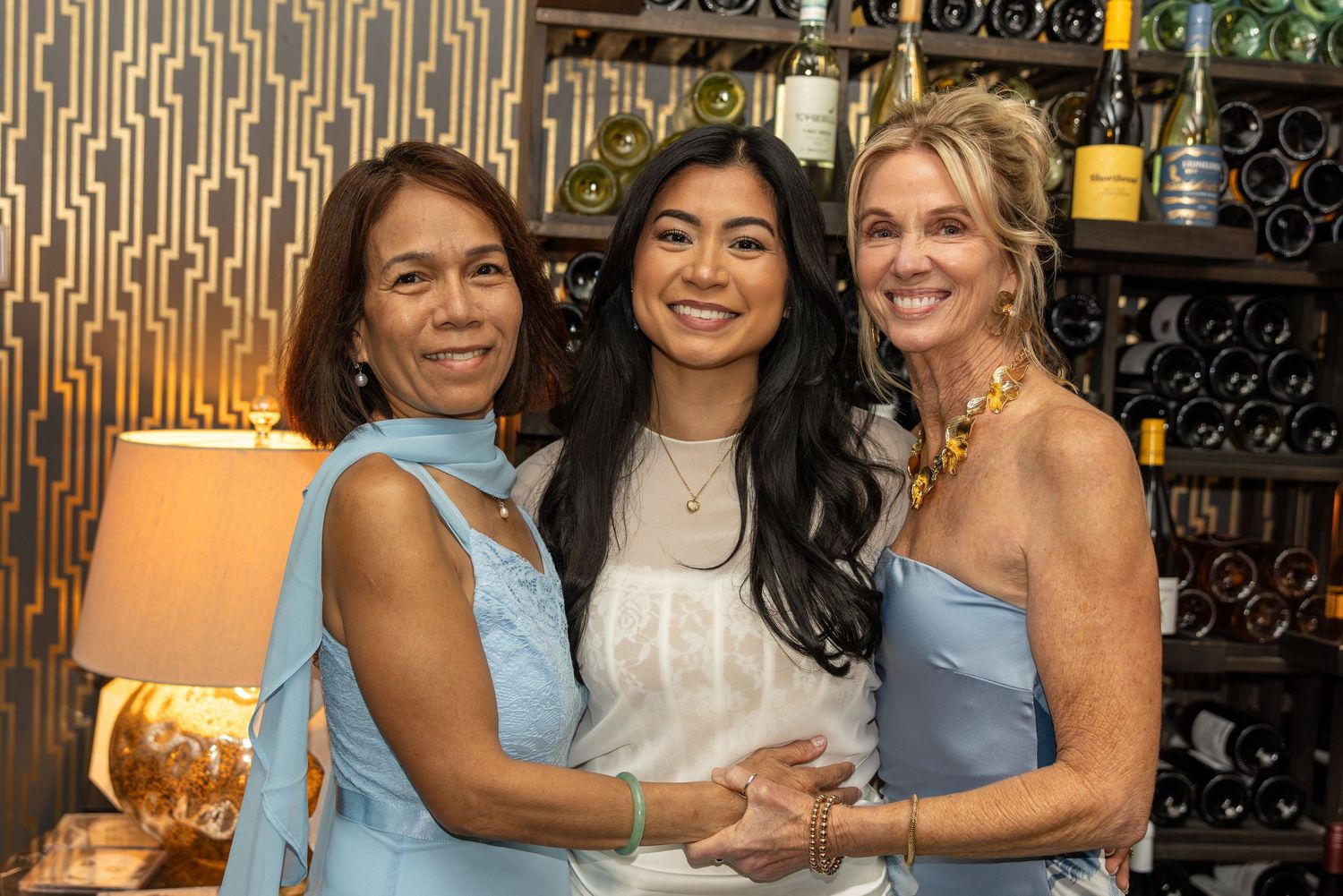 Three women smiling together at a wine bar, wearing blue and white outfits at a social gathering.