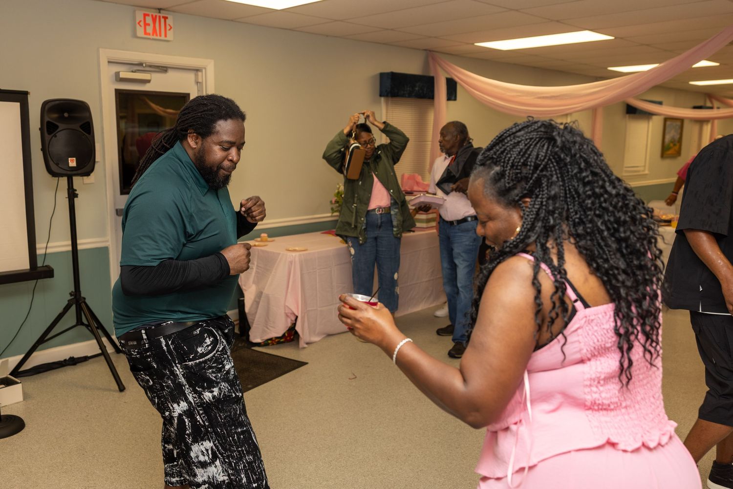 Two people dancing at an indoor party event with guests socializing in the background near decorated tables.