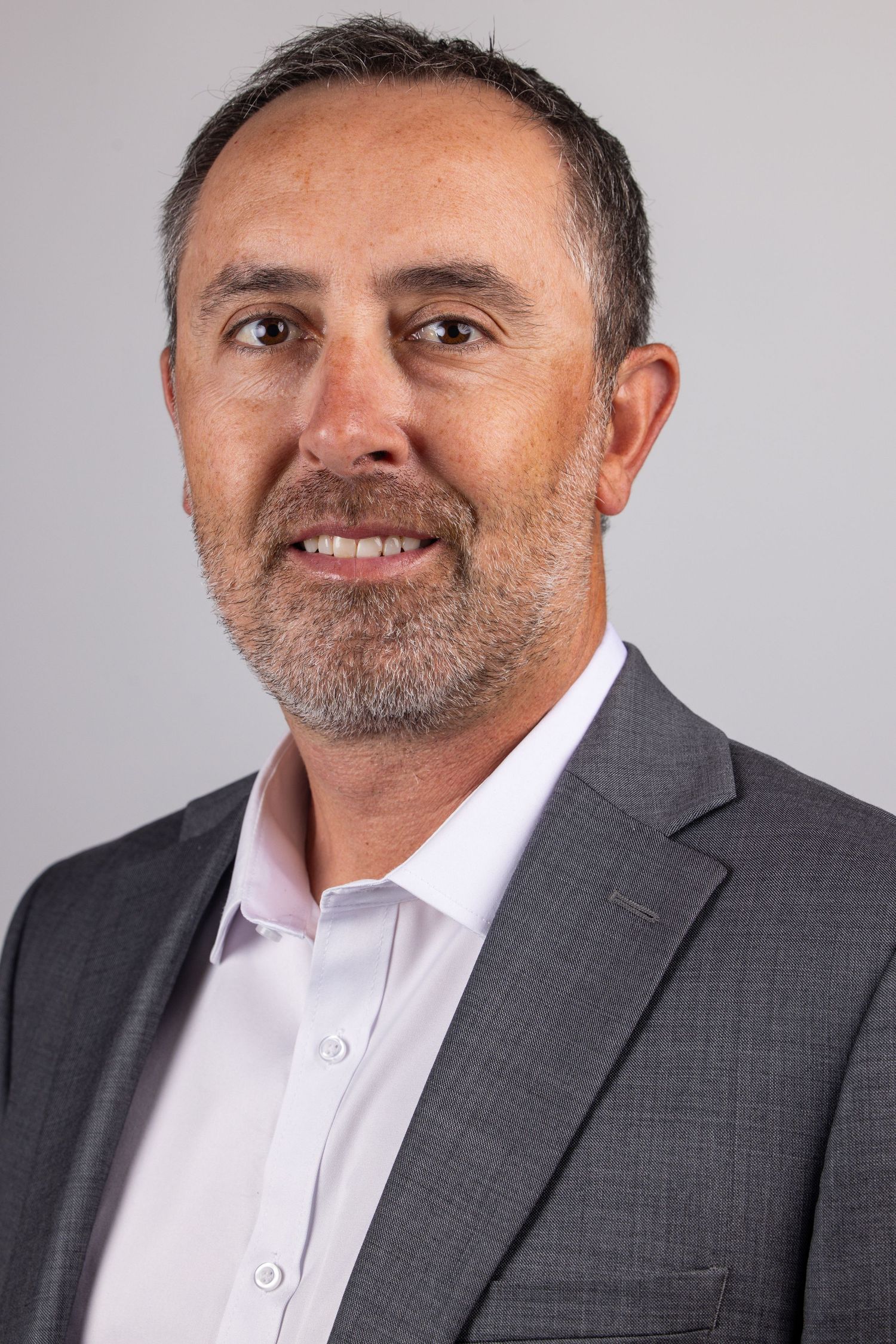 Professional headshot of a smiling middle-aged man with a beard, wearing a gray blazer and white shirt.