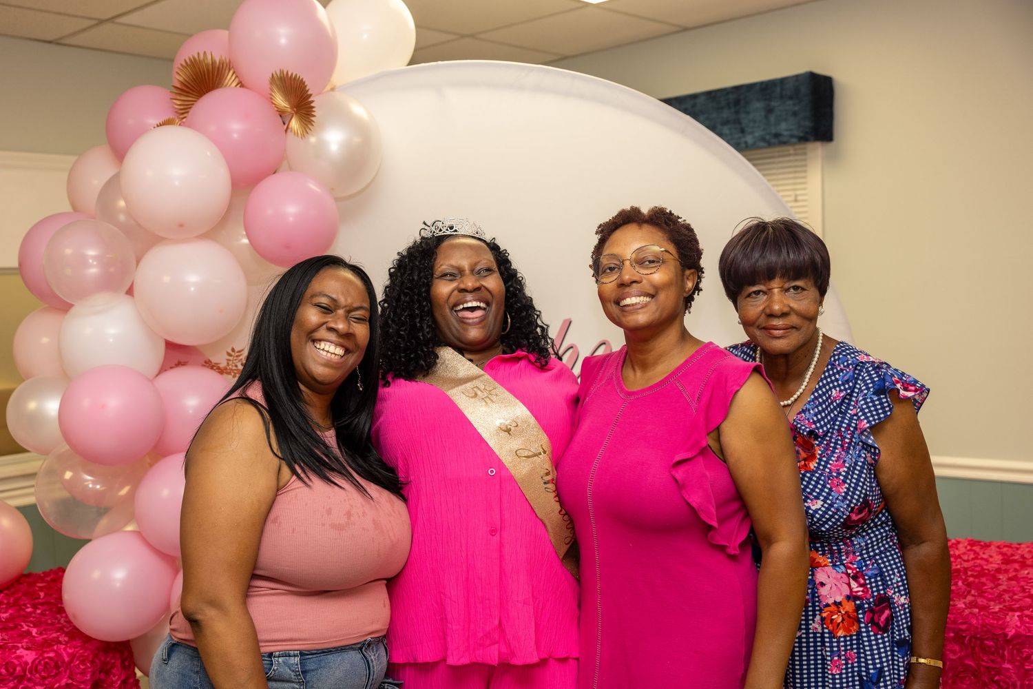 Four women in pink outfits smile together at a celebration with pink and white balloon decorations.