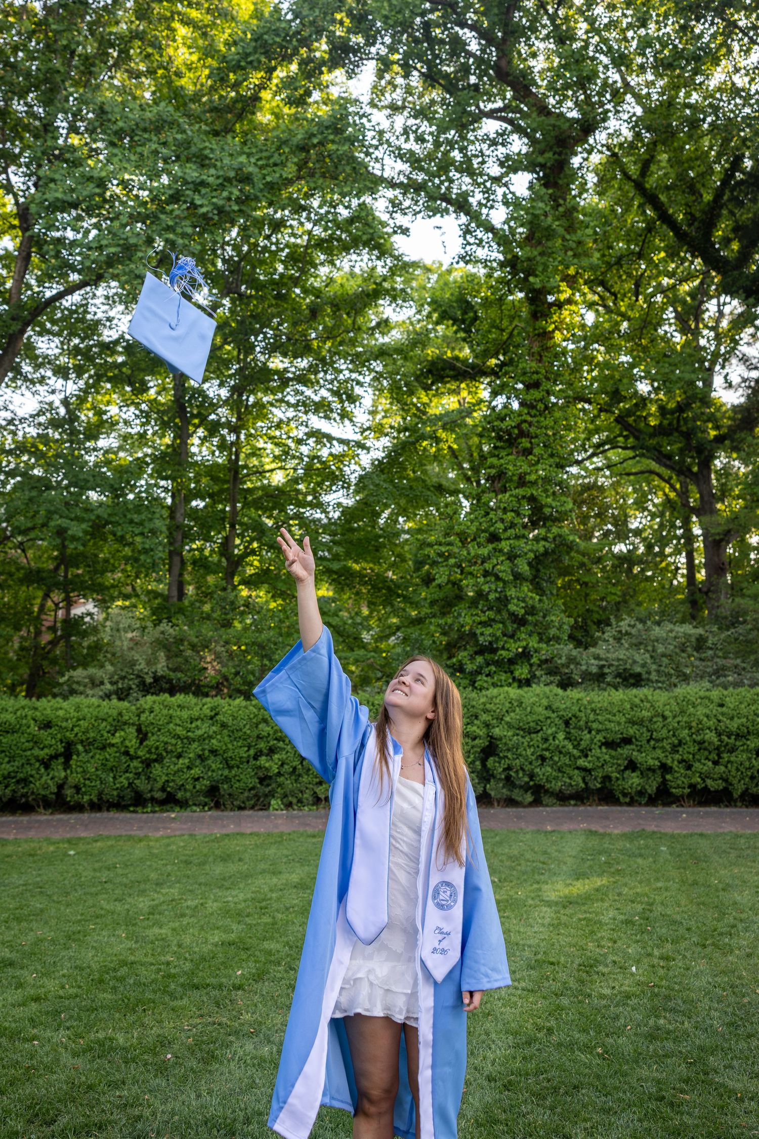Young woman in light blue graduation gown tossing cap in the air outdoors among lush green trees.