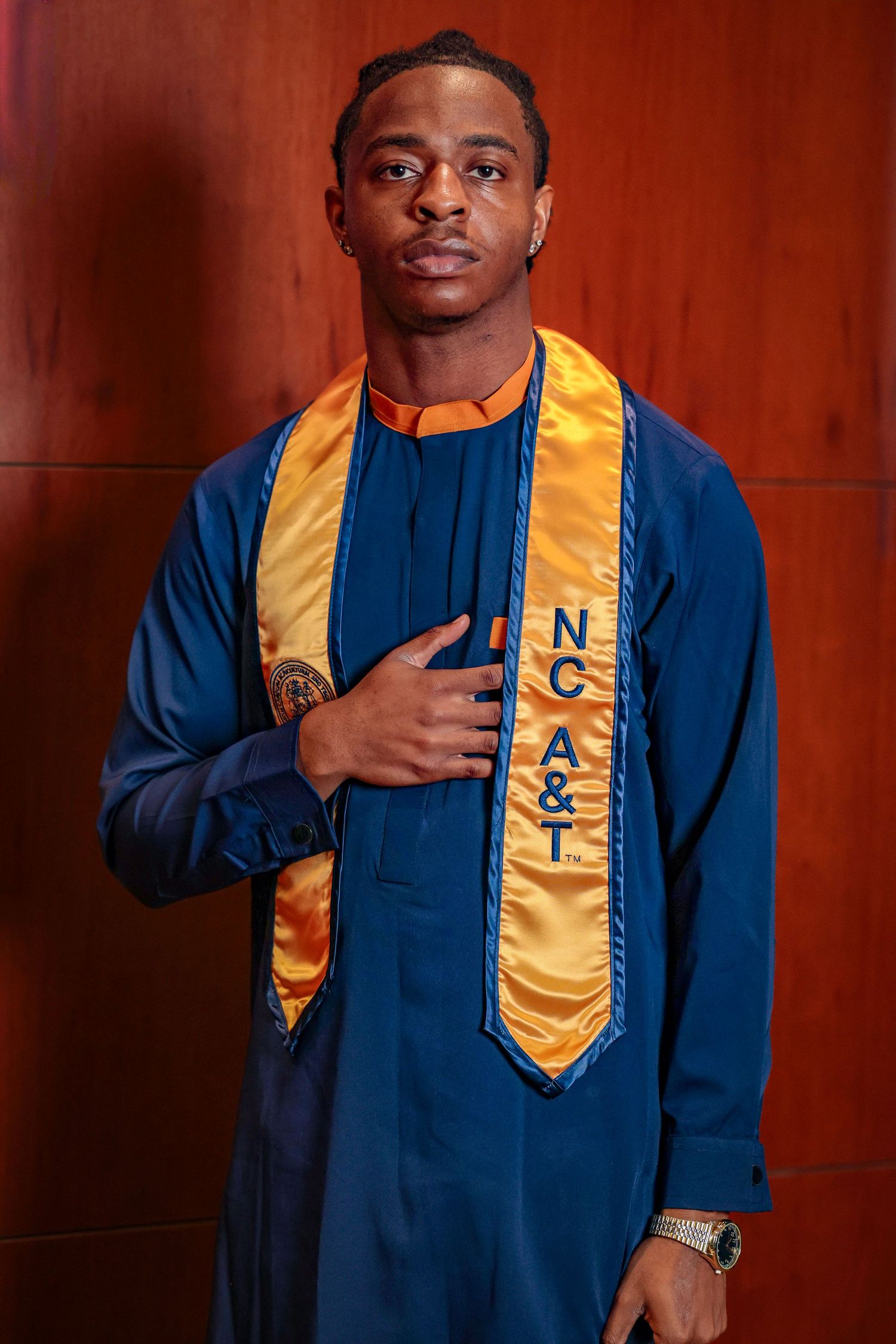 Graduate wearing blue academic regalia with gold stole poses against red wall in sequence of photos.