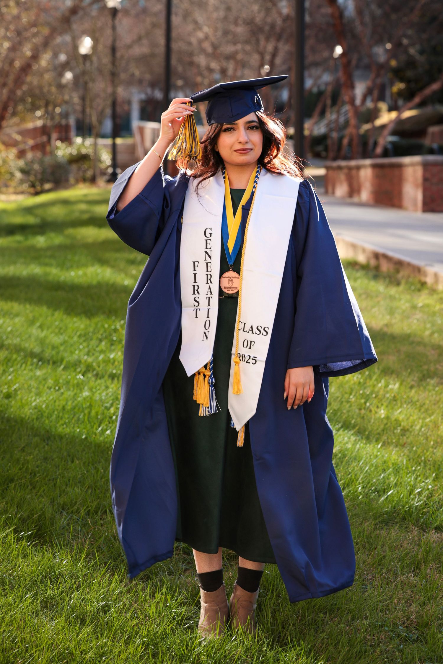 Graduate wearing navy blue academic regalia poses outdoors on a grassy hill during graduation season.