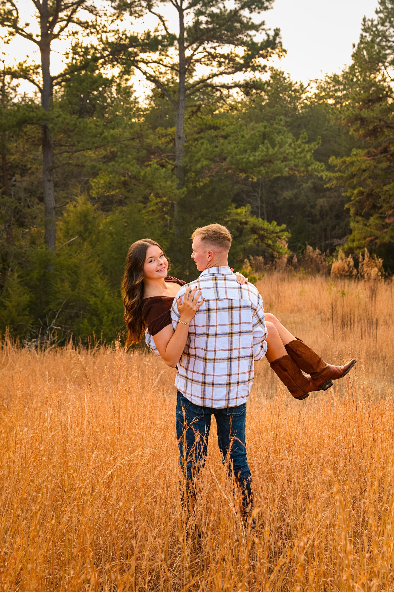 Playful couple dancing in golden field during sunset with pine trees in background.
