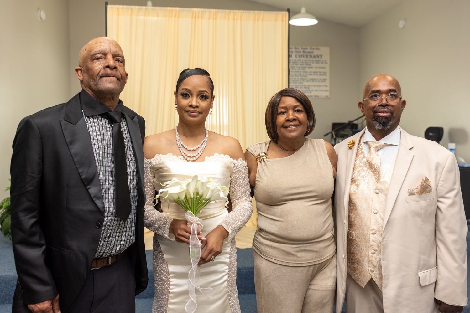 A bride in a white gown holds flowers, posing with three guests at a wedding celebration indoors.