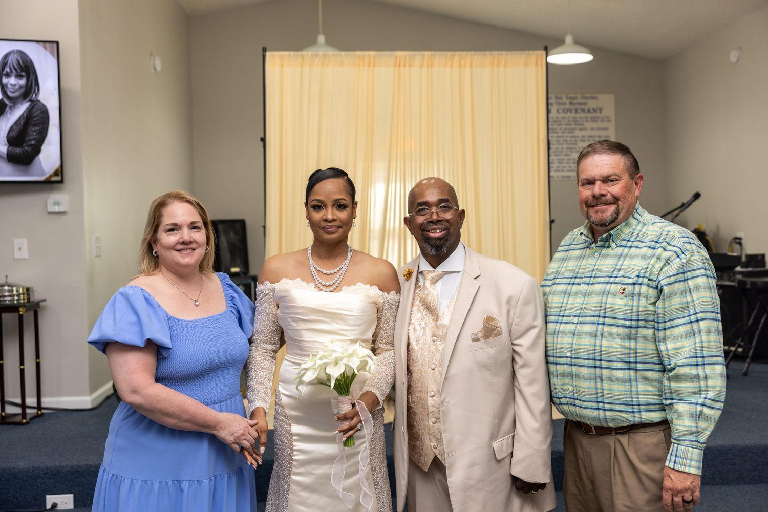 Bride and groom pose with two guests at a wedding ceremony inside a decorated venue.