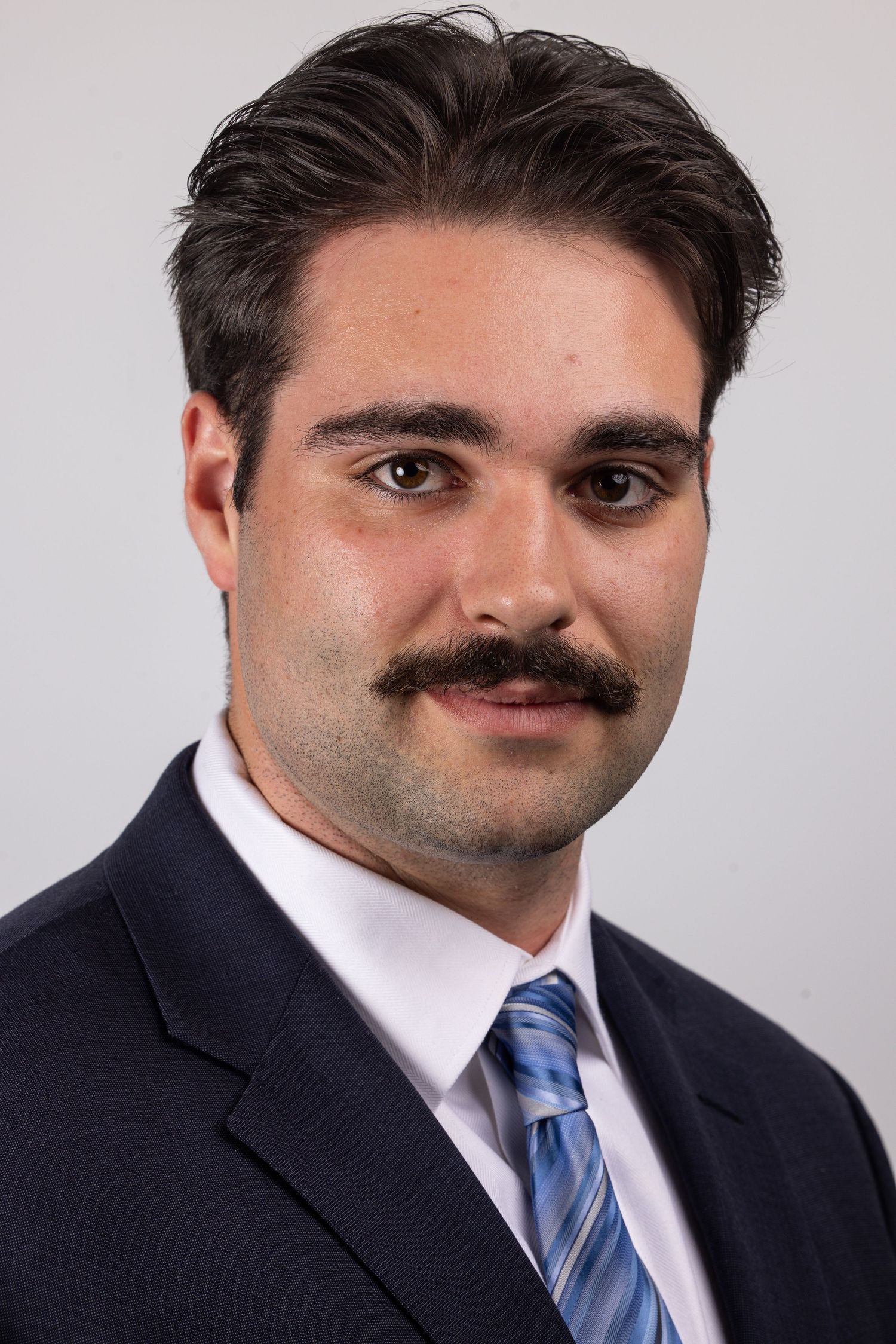 Professional headshot of a young man with dark hair and mustache wearing a navy suit and blue striped tie.