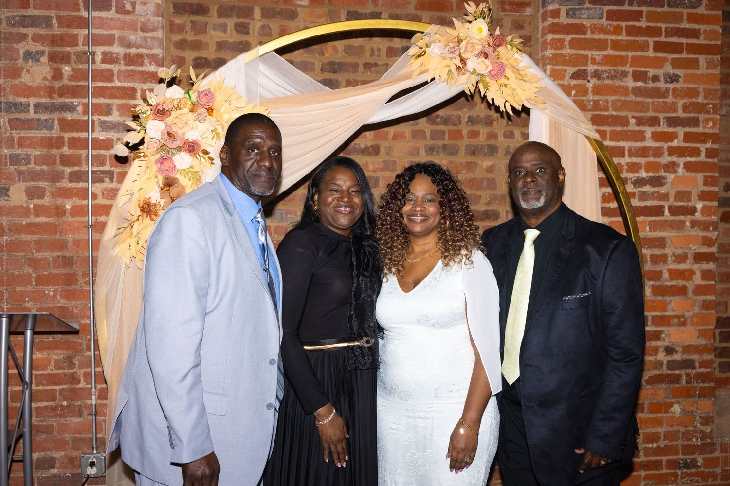 Four people smiling together under a floral arch at a wedding celebration in a brick-walled venue.