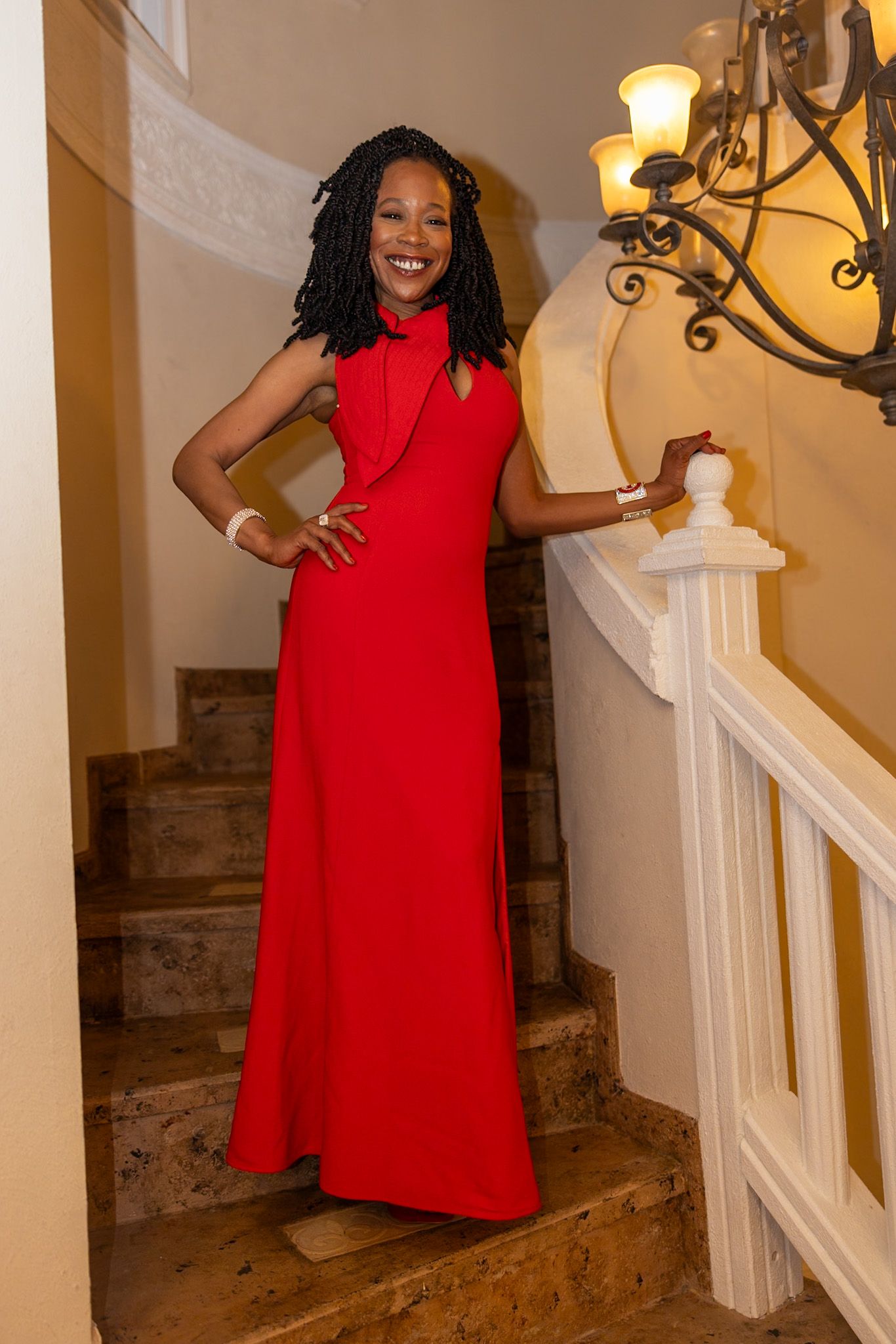 A person in a bright red dress poses playfully near a white staircase with warm lighting in an indoor setting.