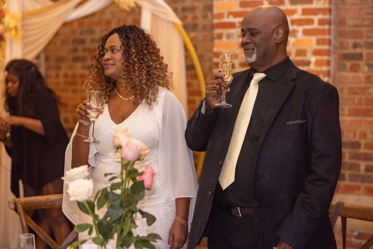 A couple in wedding attire toasts with champagne glasses at their elegant brick-walled wedding reception.