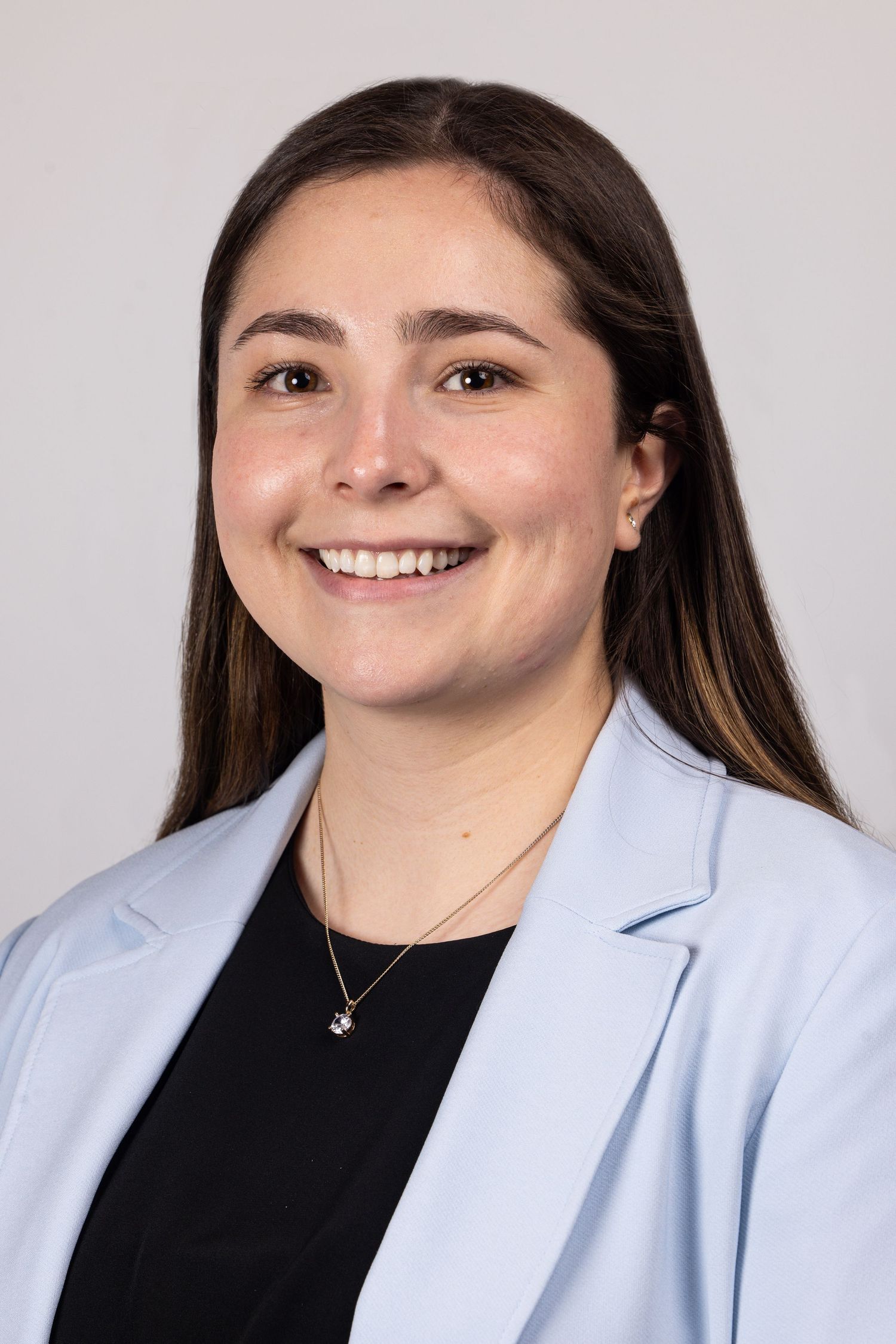 Professional headshot of a smiling young woman with long brown hair wearing a light blue blazer and gold necklace.