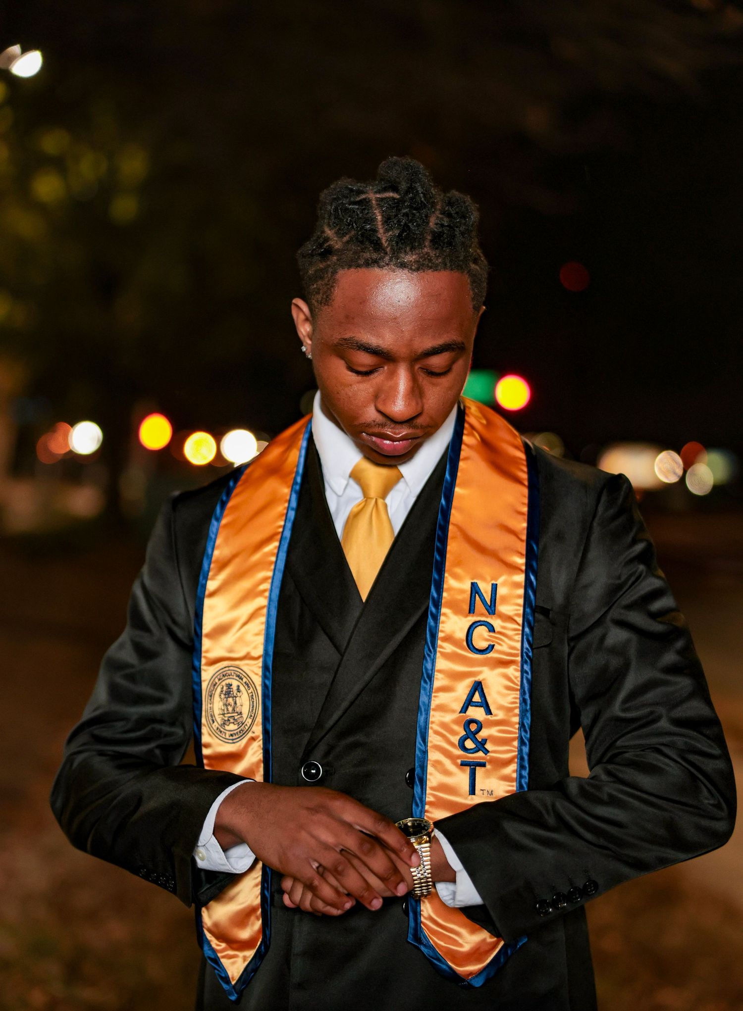 Graduate wearing black suit and gold stoles smiles during evening graduation photoshoot.