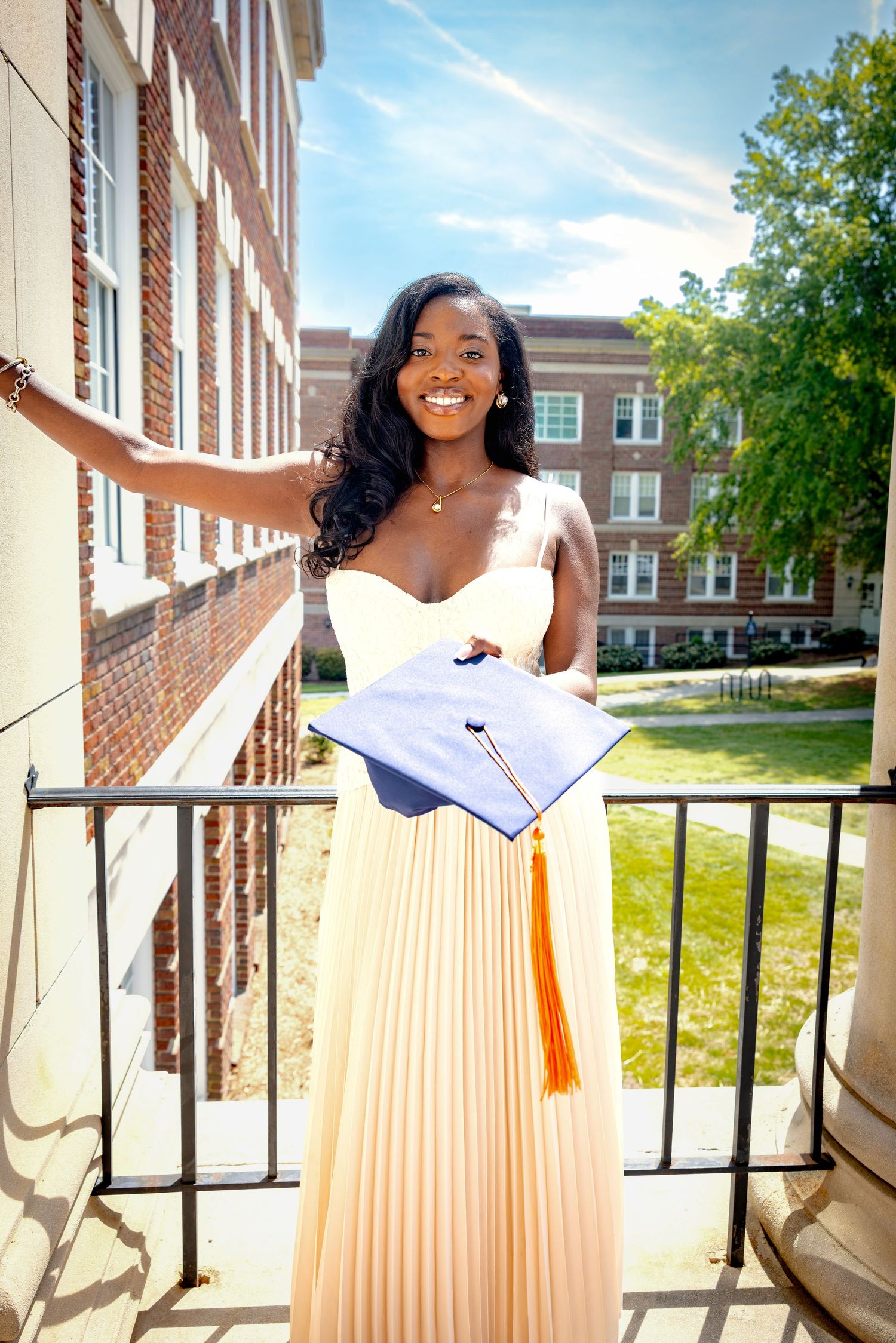 Smiling young woman in elegant cream pleated gown holding graduation cap with orange tassel on university building balcony.