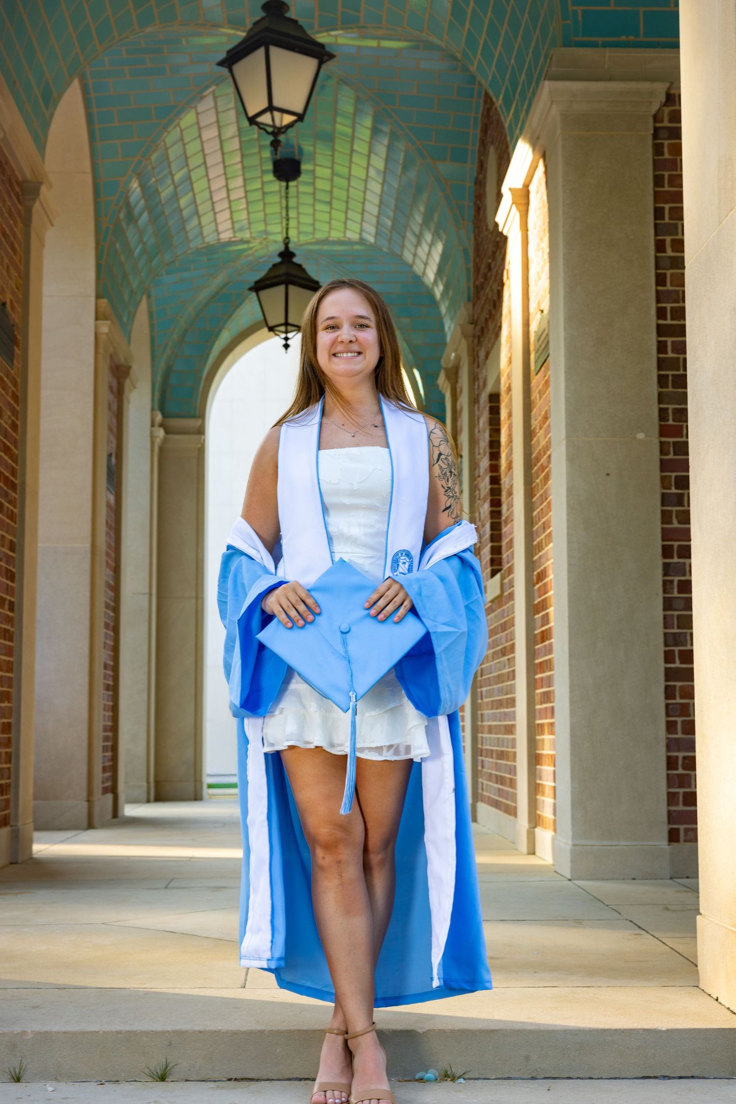 Young woman in light blue graduation gown and cap smiling in a brick archway corridor on graduation day.