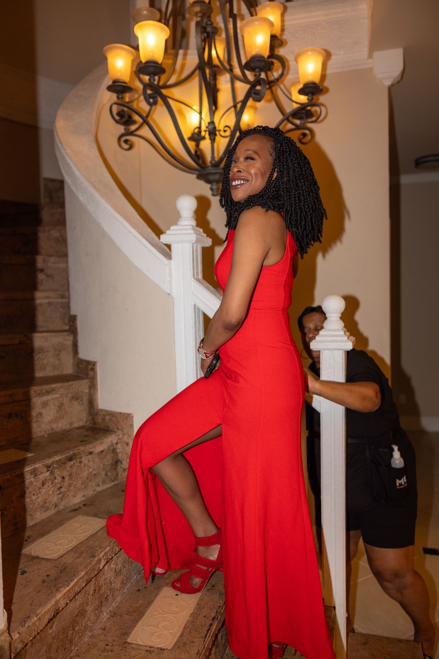 A person in a flowing red dress poses playfully on an elegant staircase with ornate lighting fixtures above.