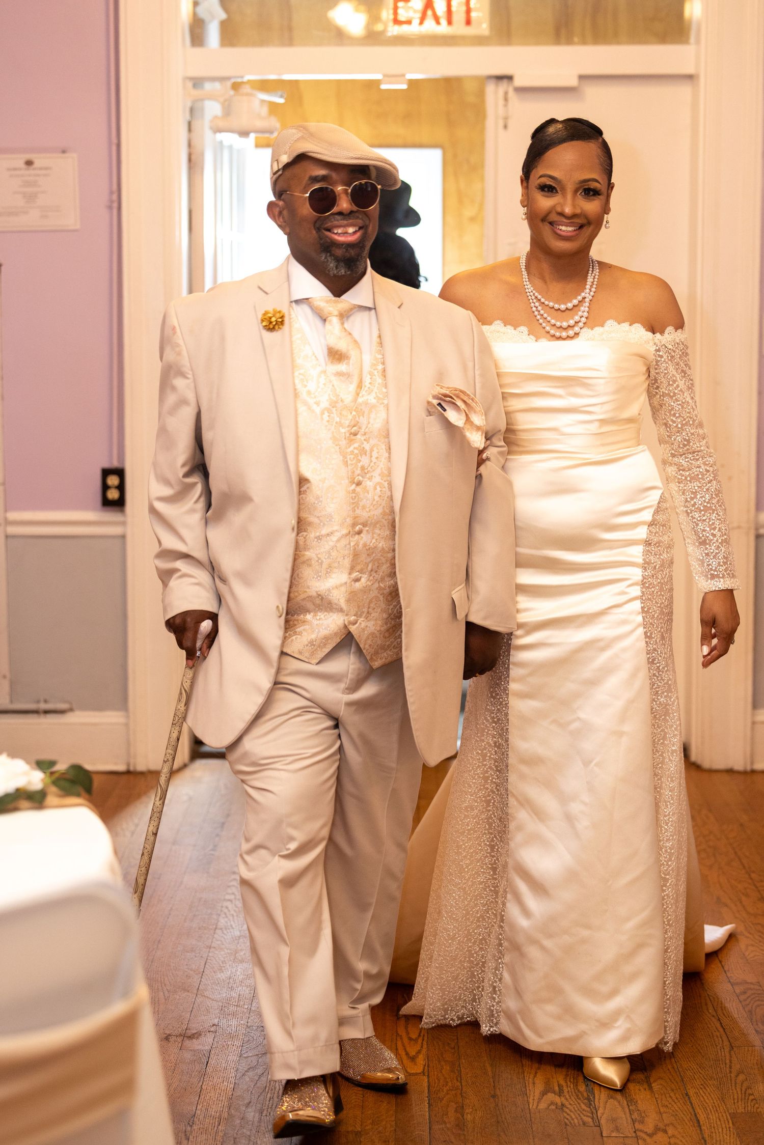 Elegant couple in matching ivory wedding attire walking together, groom with cane and hat, bride in satin gown with pearls.