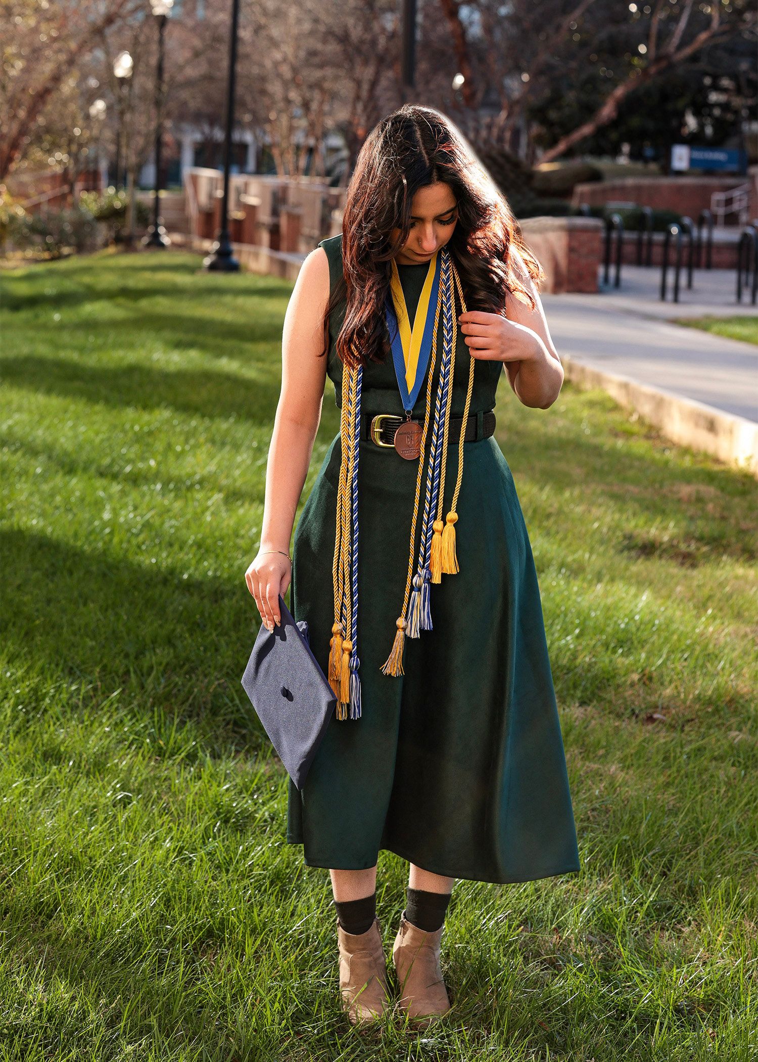 Graduate in a dark green dress stands on a grassy hill during a graduation photoshoot.