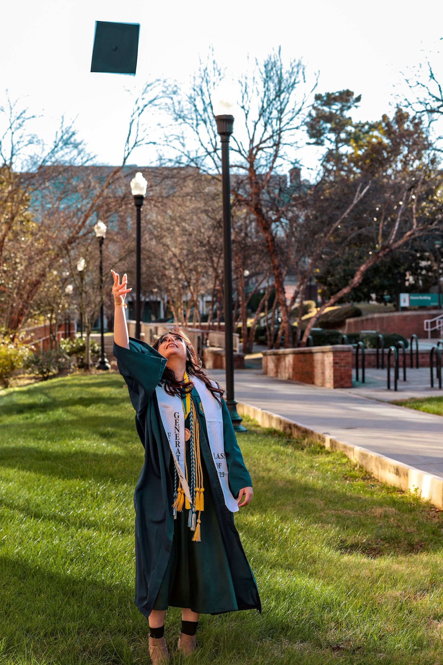 Graduate celebrates on campus lawn wearing traditional blue graduation cap and gown with ceremonial stoles.