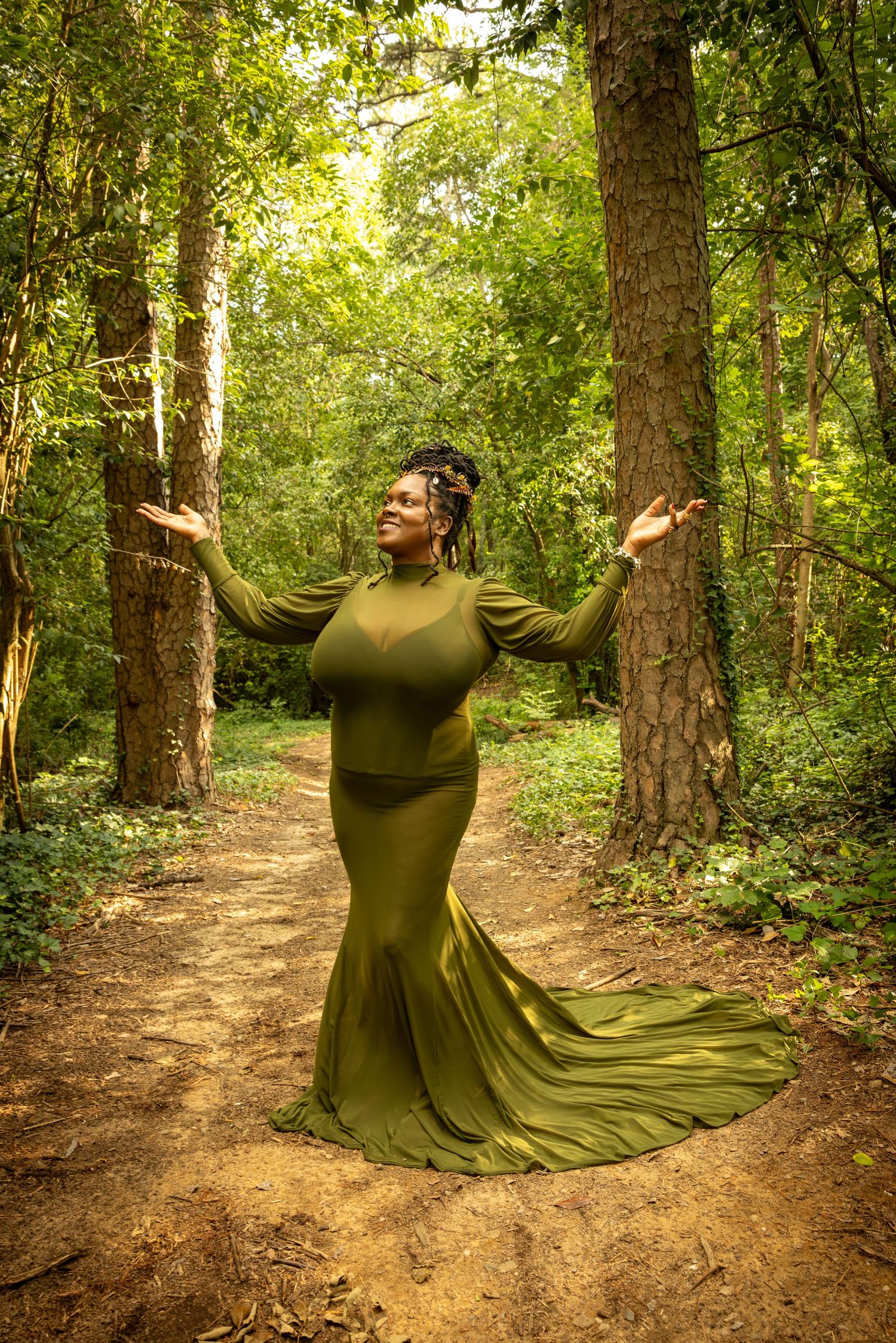 Woman in olive green gown poses gracefully on forest path with arms raised among tall trees.