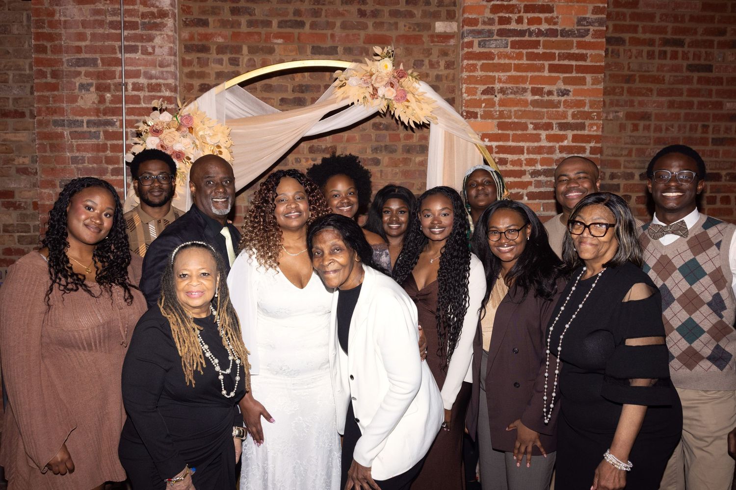 Group of elegantly dressed people posing together at a wedding reception near a floral arch against a brick wall.