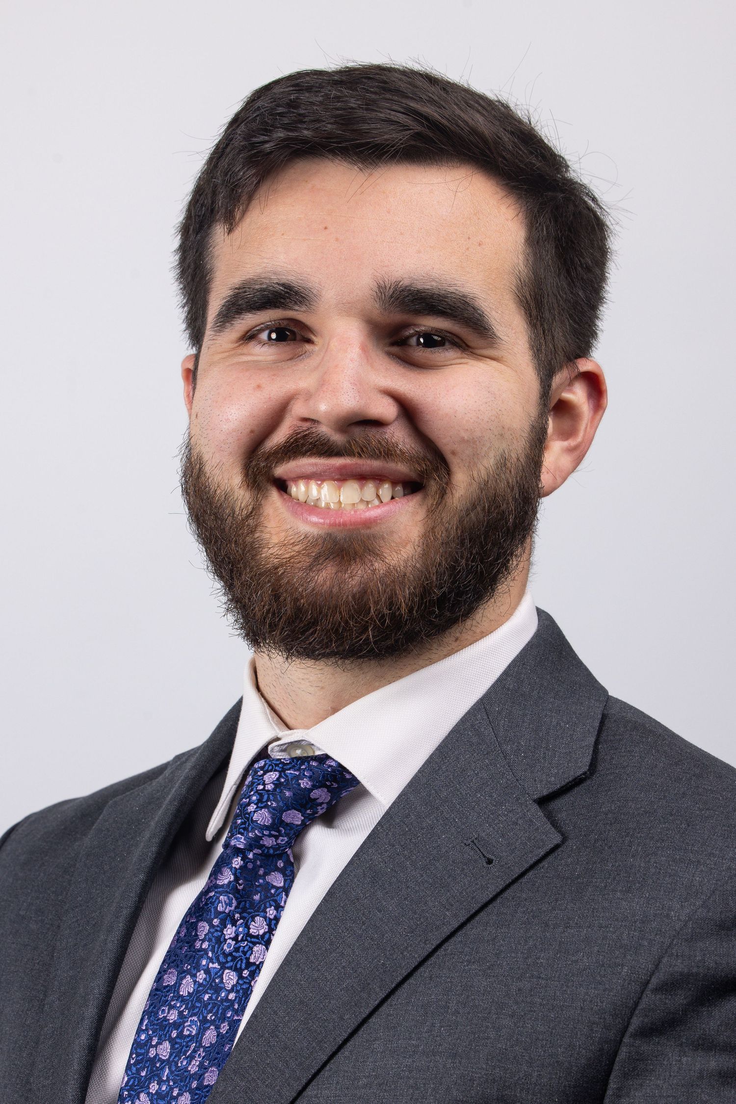 Professional headshot of a young bearded man in a gray suit with a blue floral tie.