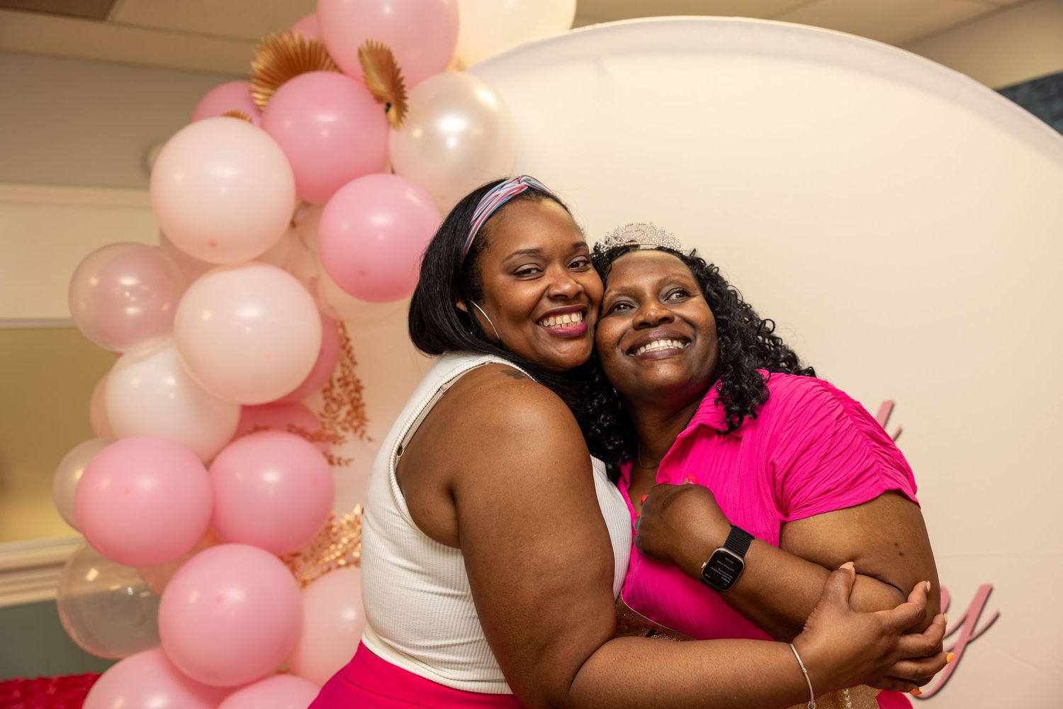 Two women smiling and hugging at a pink balloon baby shower celebration.