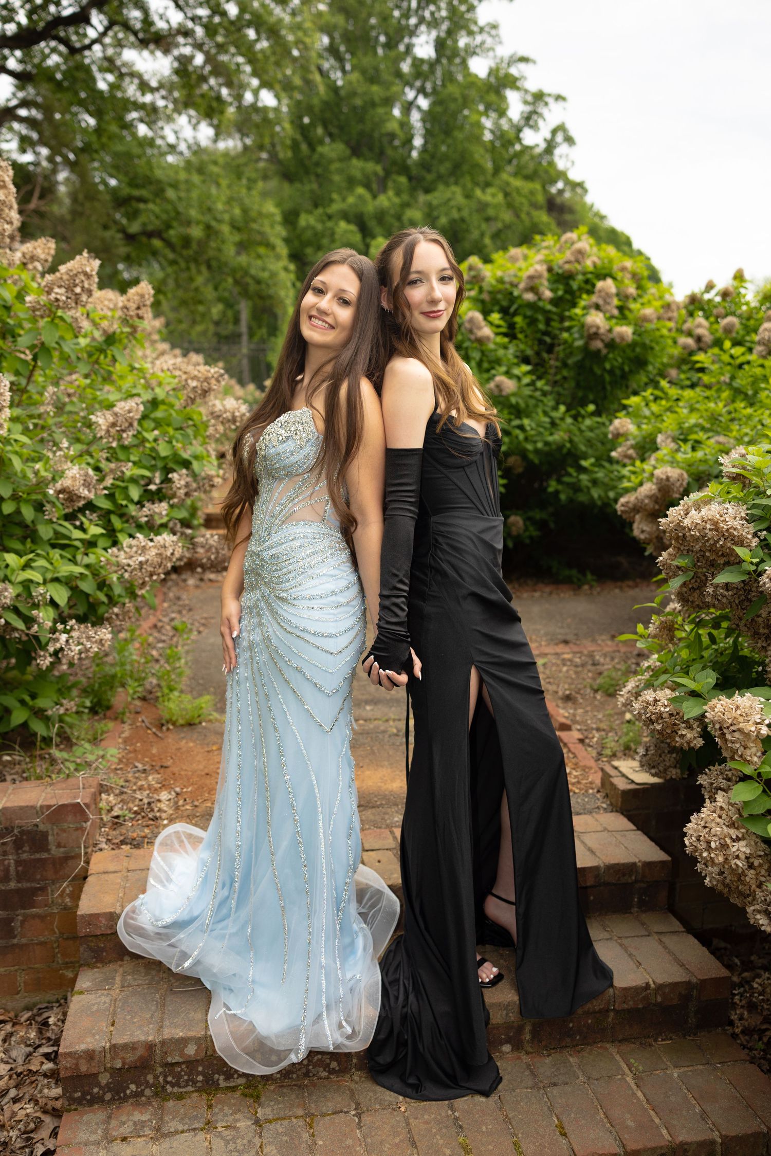 Two young women in elegant prom gowns, one blue beaded and one black, pose back-to-back in a lush garden setting.