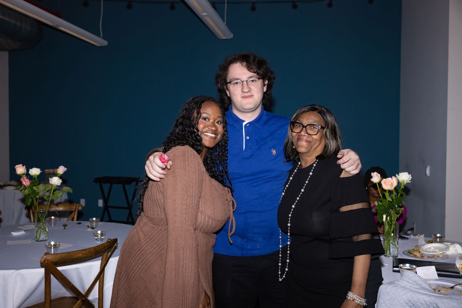 Three friends smiling and posing together at an elegant event venue with teal walls and floral table settings.