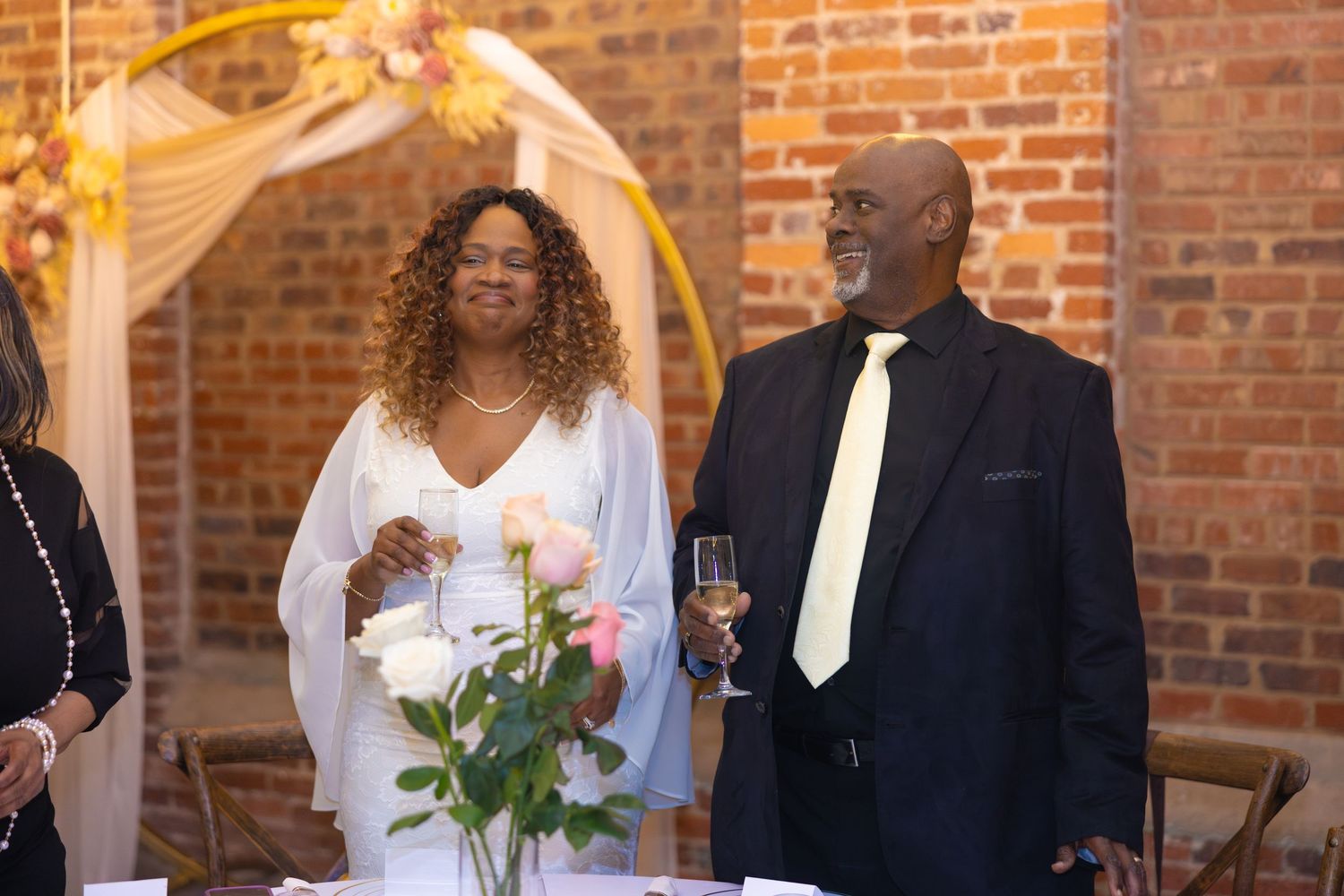 Couple smiling at wedding reception near floral arch, man in black suit with tie holding champagne glass.