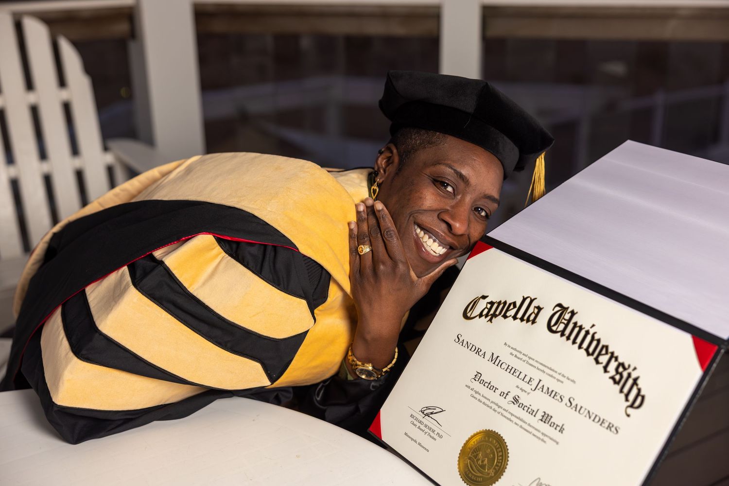 Graduate in yellow and black regalia proudly poses with Capella University Doctor of Social Work diploma.