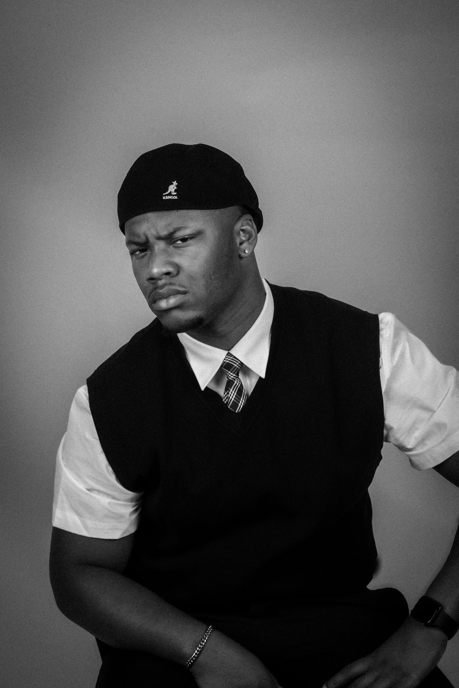 Black and white portrait of a young man wearing a Kangol cap and sweater vest with a tie.