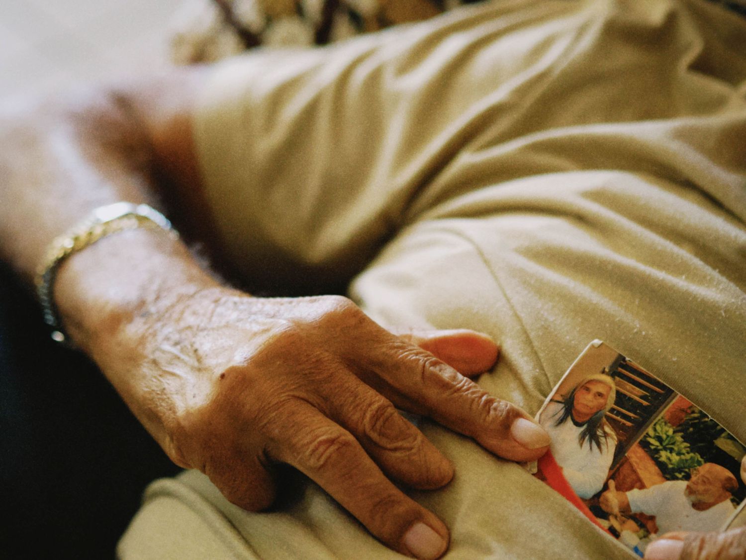 Close-up of an elderly person's wrinkled hand resting on lap, wearing a bracelet, holding a small family photo.
