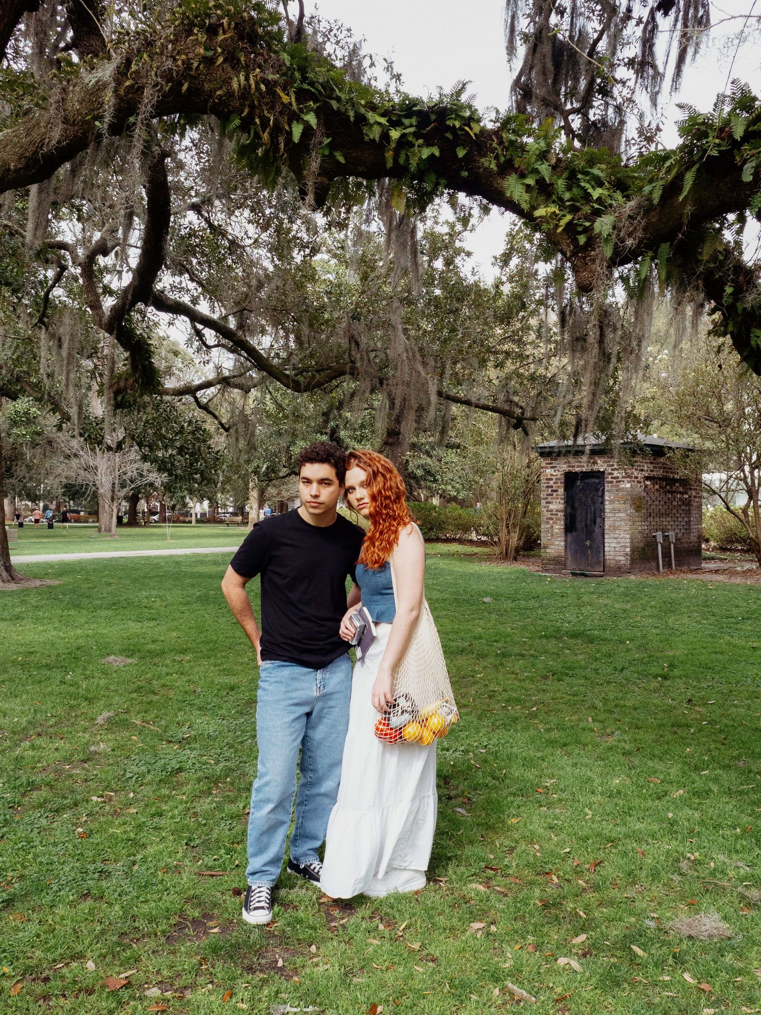 A couple shares a tender moment on a grassy lawn surrounded by Spanish moss-draped trees in a romantic outdoor setting.