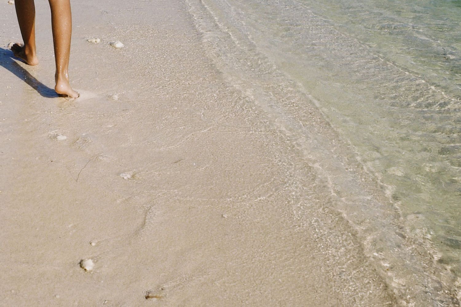 Person standing barefoot on white sandy beach with clear turquoise water washing ashore.
