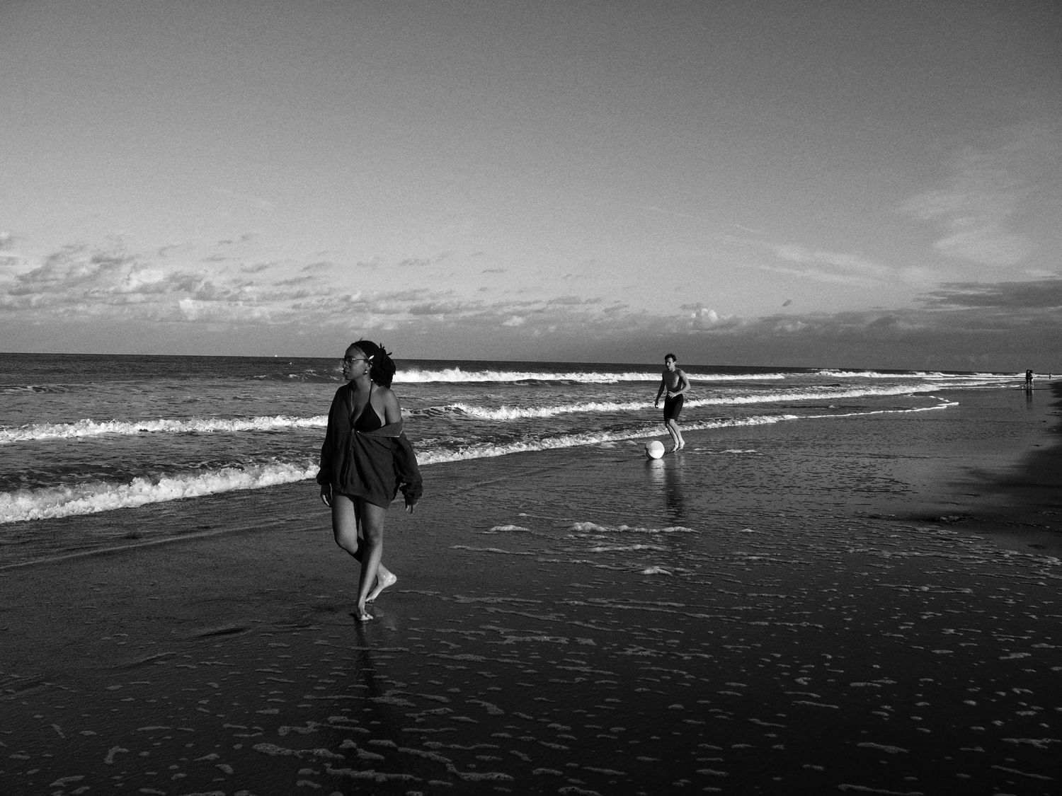A surfer walks along a dark sandy beach carrying their surfboard against a dramatic black and white ocean backdrop.