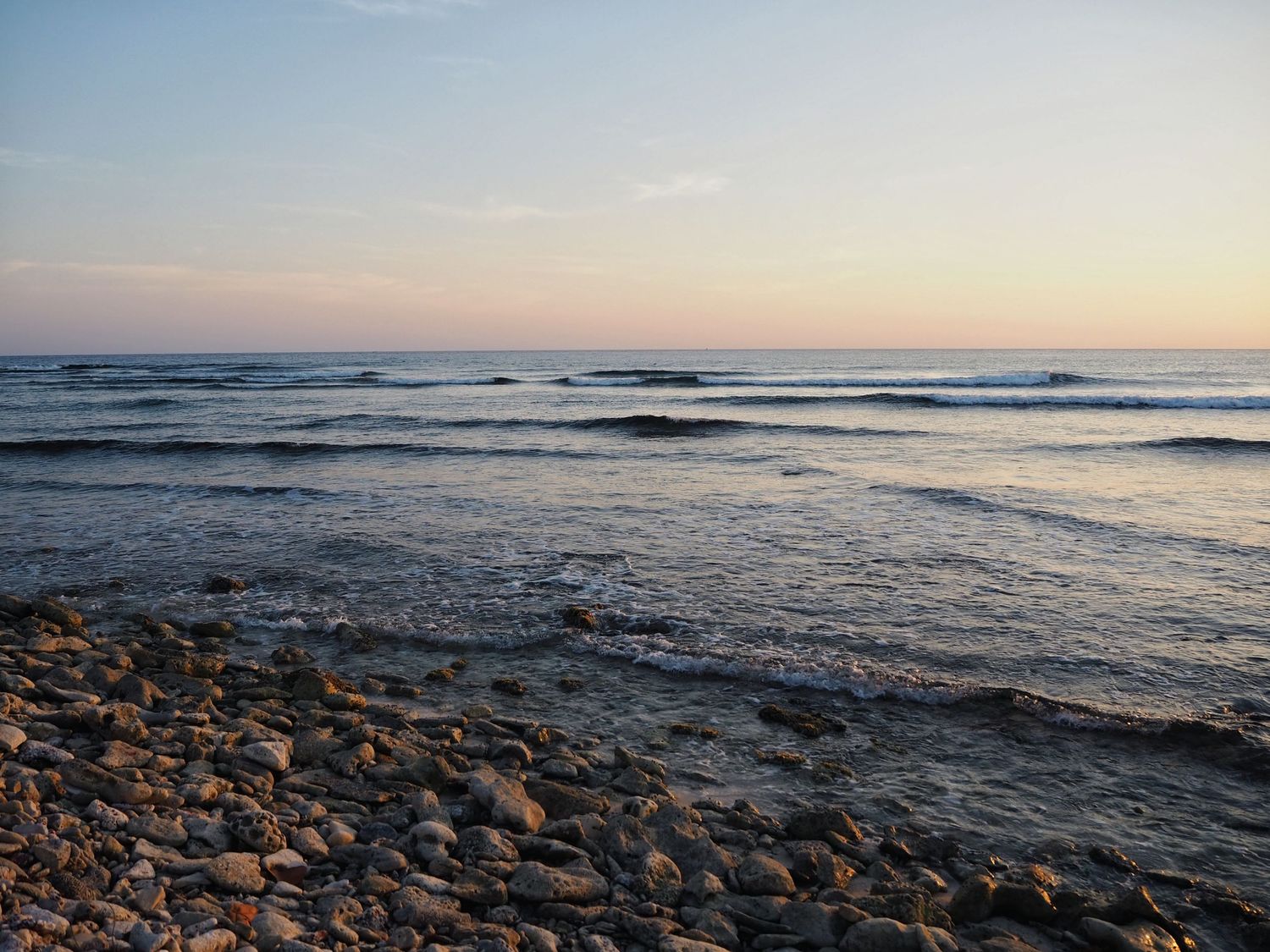 Tranquil ocean waves roll onto a rocky beach at sunset with warm golden light reflecting on the water's surface.